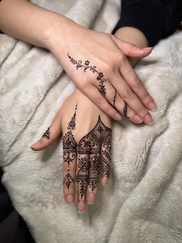 Two hands with intricate black henna tattoos resting on a soft, beige blanket. One hand has a floral and vine design on the wrist, while the other has detailed geometric and floral patterns covering the back of the hand and fingers.