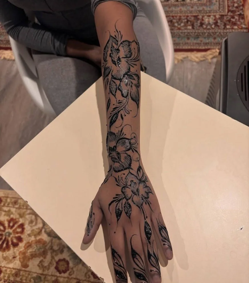 A person's arm and hand covered with a black floral henna tattoo design, resting on a light-colored table. The background includes a patterned rug and part of a chair.