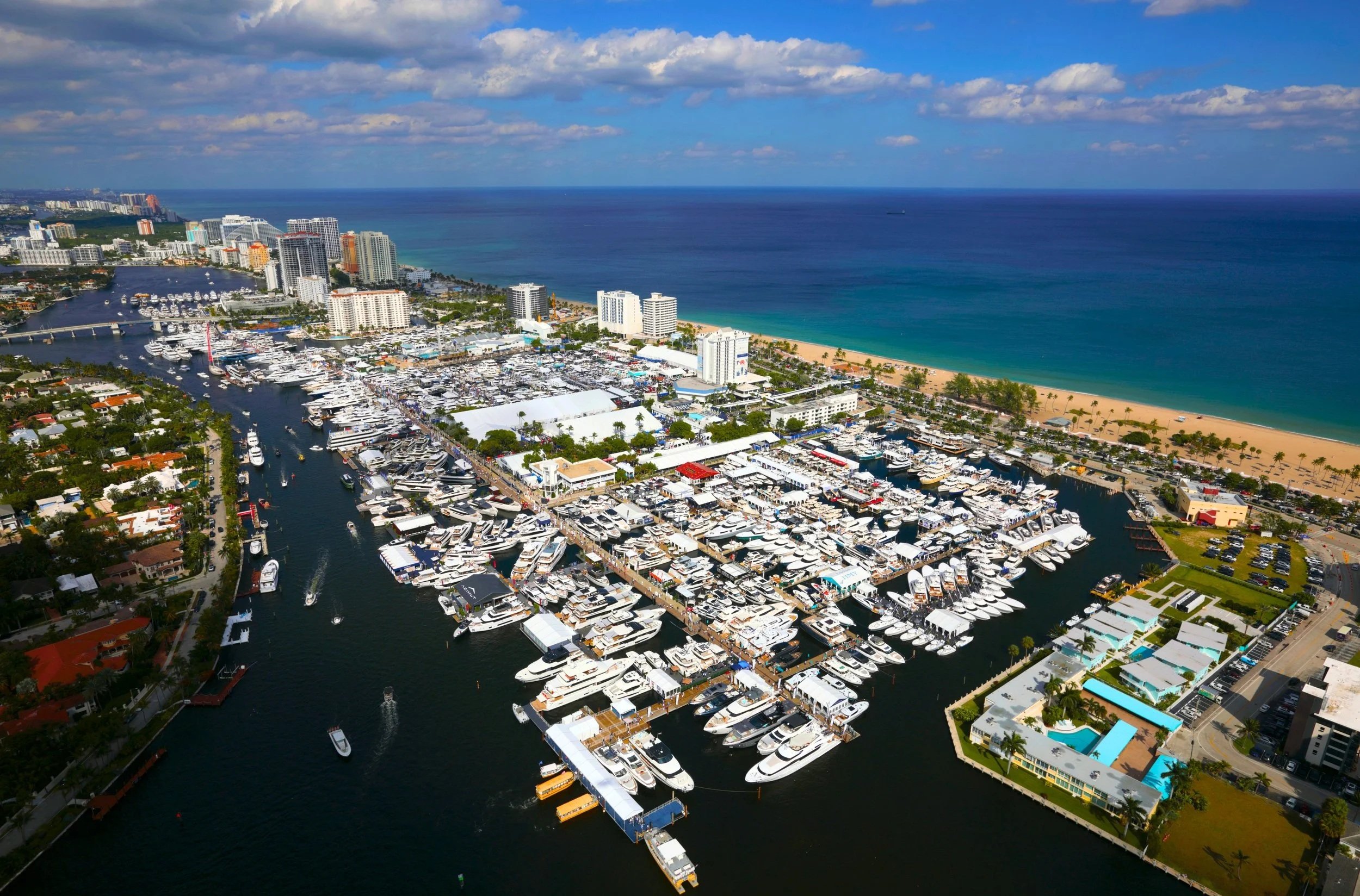 Aerial view of a Fort Lauderdale Boat Show with numerous yachts and boats, high-rise buildings along the coast, beach with palm trees, and the ocean under a partly cloudy sky.