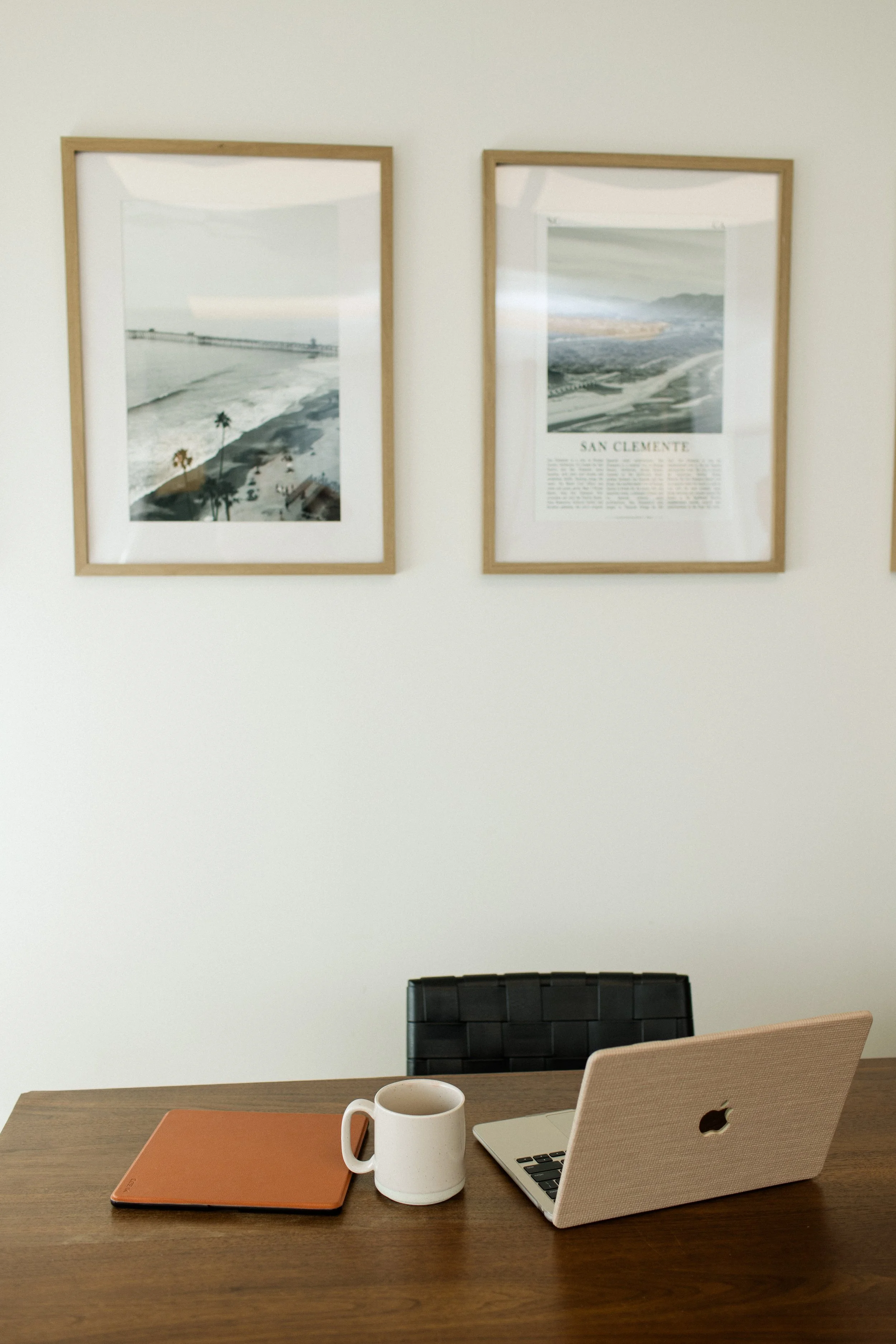 A desk with a closed orange notebook, a white coffee mug, and an open beige laptop, with a black and a beige chair behind it. On the wall above are three framed photographs of coastal scenes.