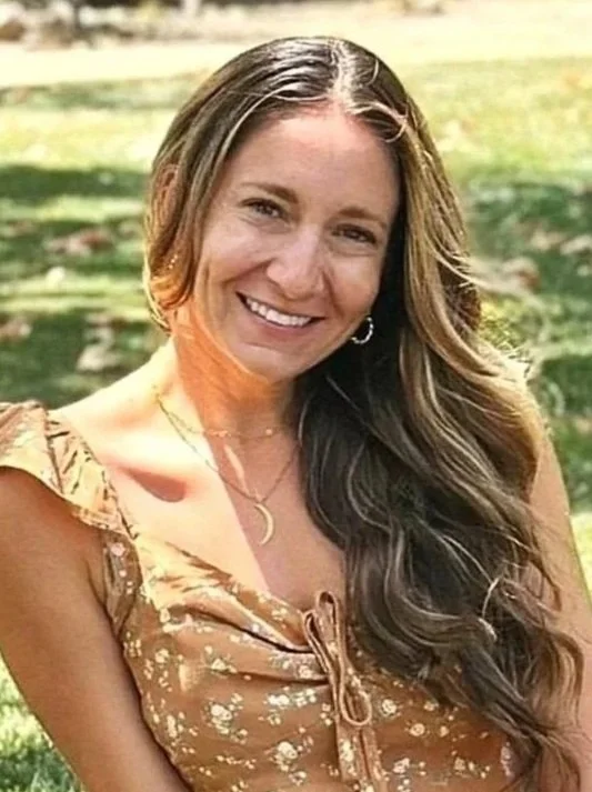 A woman with long wavy brown hair smiling outdoors on a sunny day, wearing a floral dress and layered necklaces.
