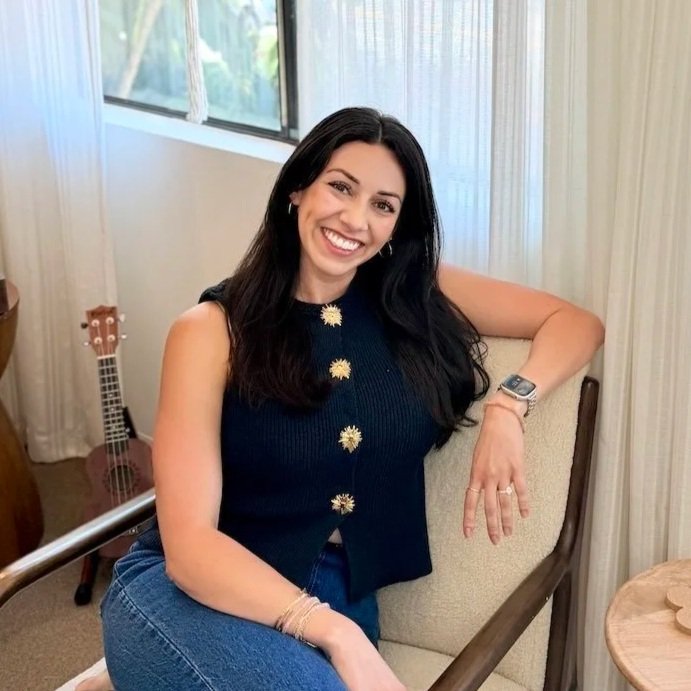 Smiling young woman with long dark hair, wearing a white dress and hoop earrings, sitting outdoors on a grassy area.