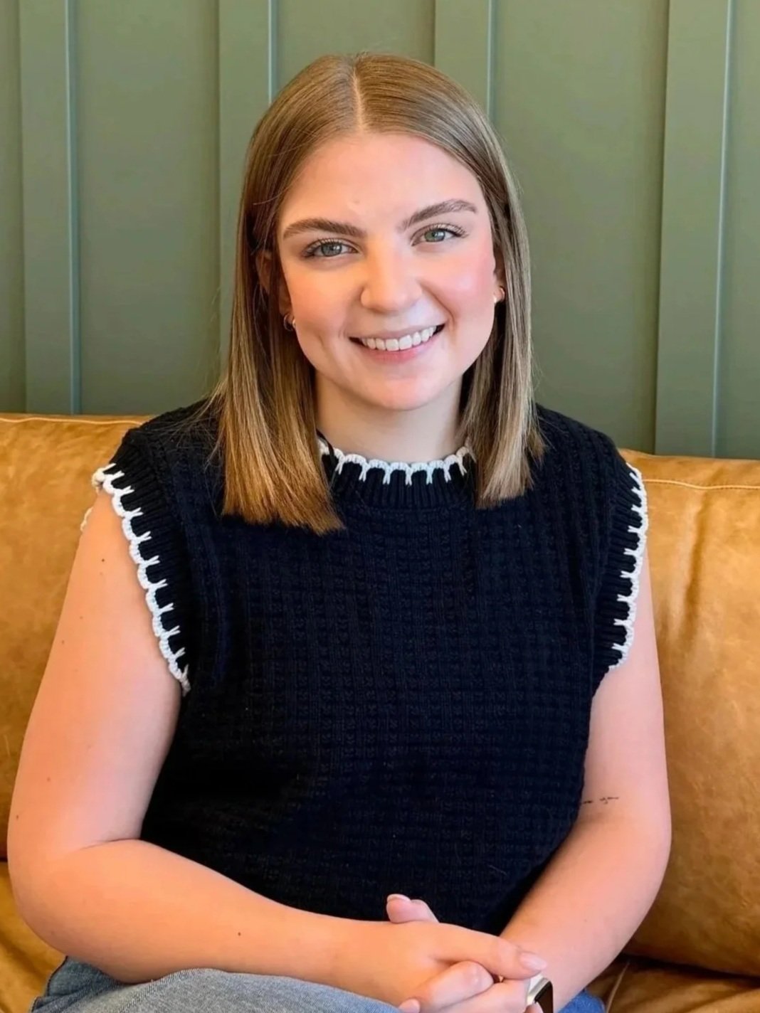 Young woman with shoulder-length light brown hair, smiling, wearing a sleeveless black top with white trim, sitting on a mustard-colored couch in front of a green paneled wall.