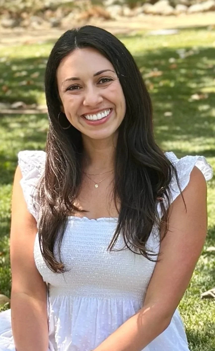 Smiling young woman with long dark hair, wearing a white dress and hoop earrings, sitting outdoors on a grassy area.