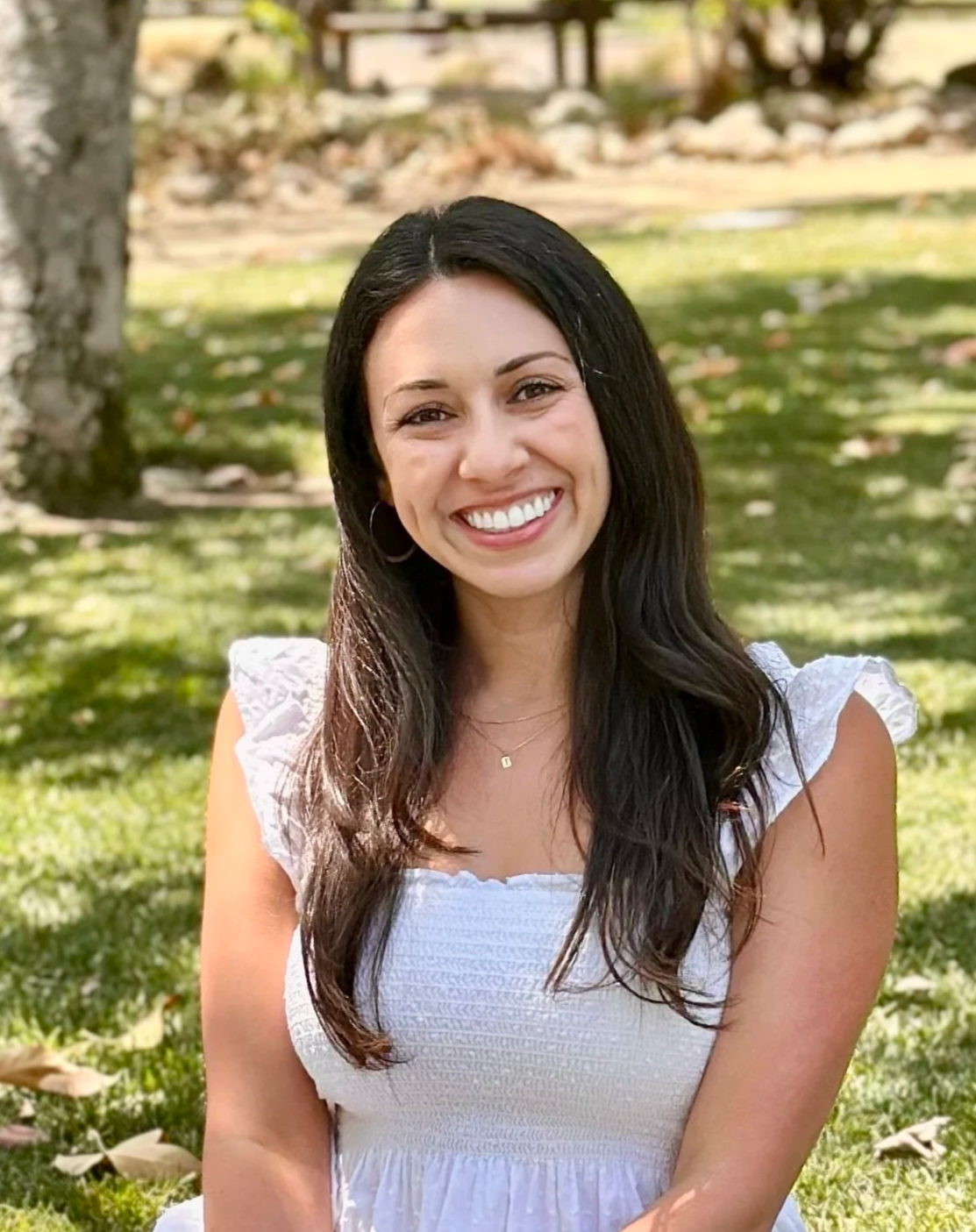 Smiling young woman with long dark hair, wearing a white dress and hoop earrings, sitting outdoors on a grassy area.