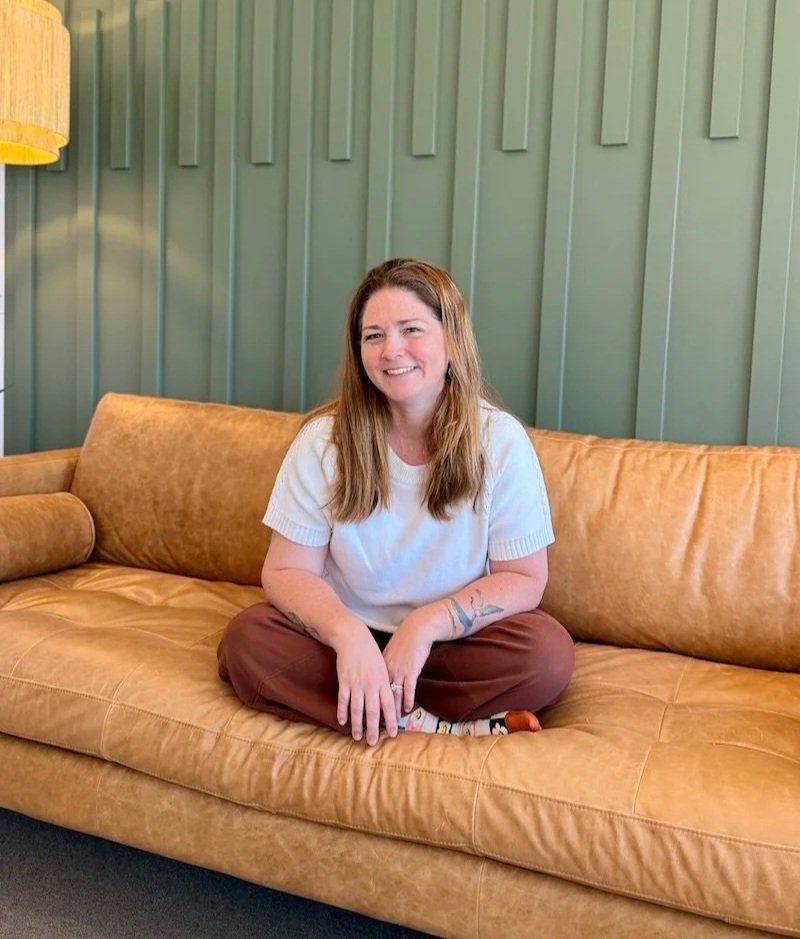 A smiling woman with shoulder-length brown hair, wearing glasses and a cream-colored dress with black and blue spots, sitting in a room with decorative shelves and a yellow patterned box in the background.