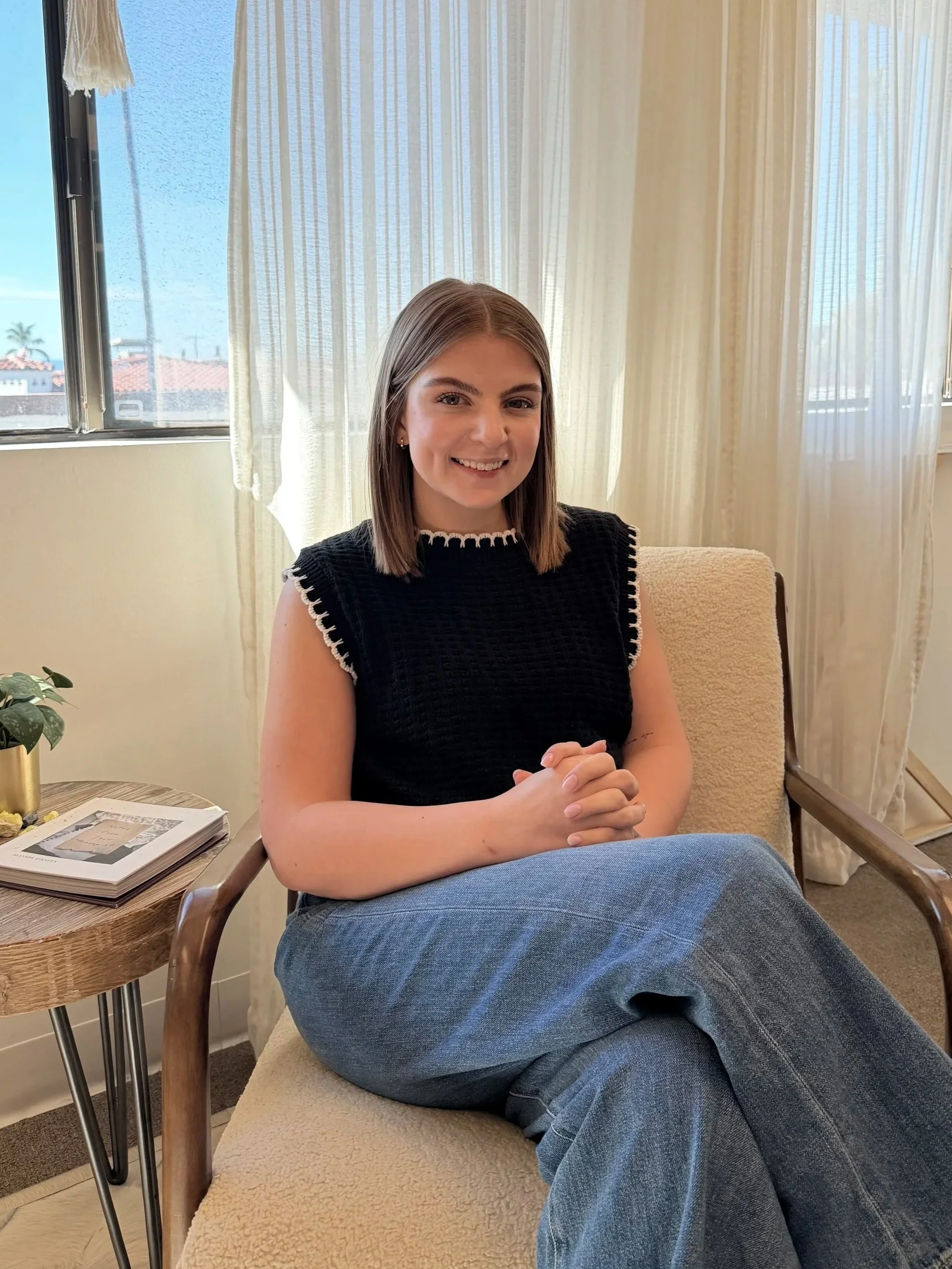 A young woman with shoulder-length brown hair, smiling, sitting on a beige armchair with her hands clasped. She's wearing a black sleeveless top with white trim and blue jeans, in a sunlit room with cream curtains and a small wooden side table with magazines and a plant.