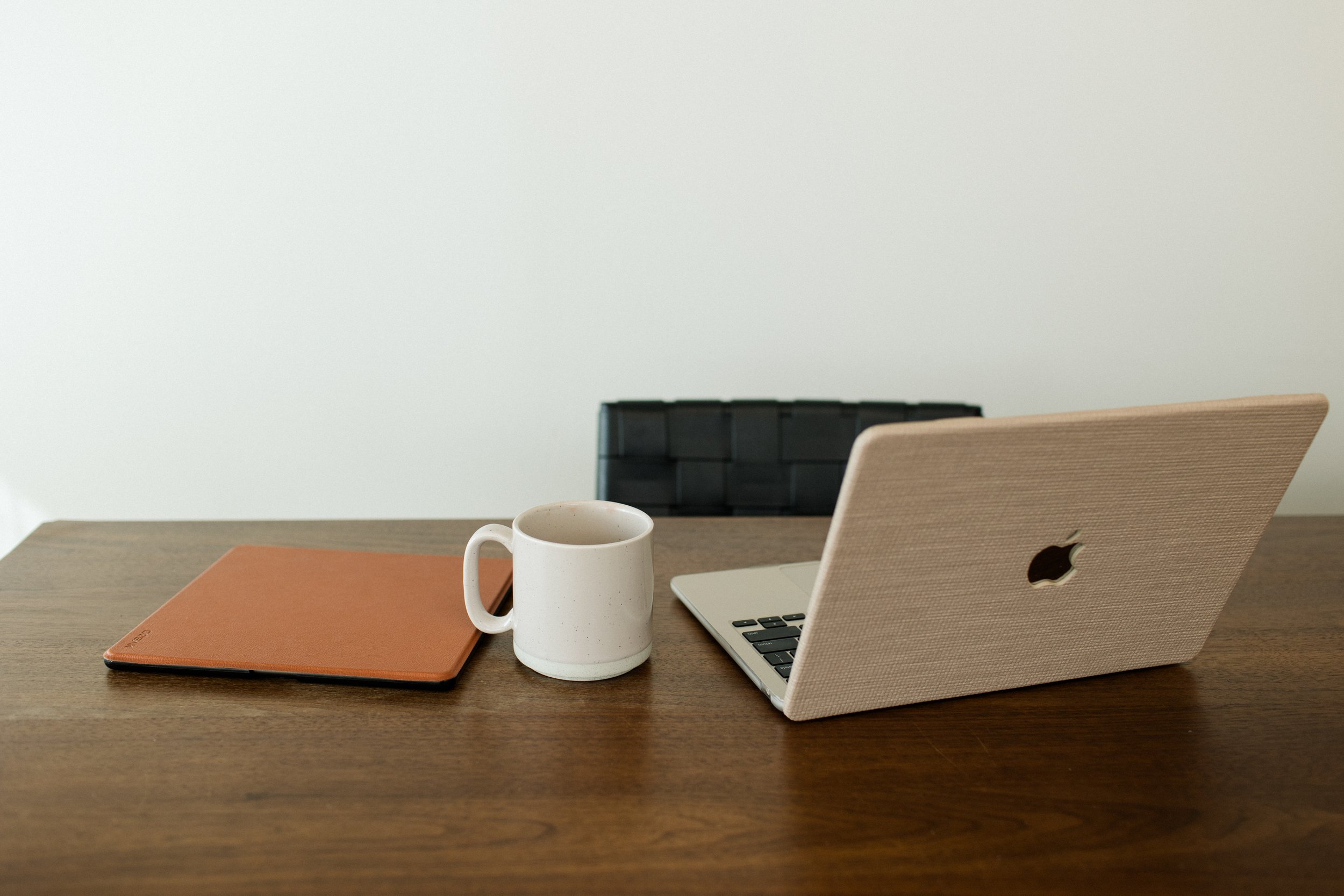 A wooden desk with a closed brown notebook, a white coffee mug, and a silver laptop with a beige fabric cover.