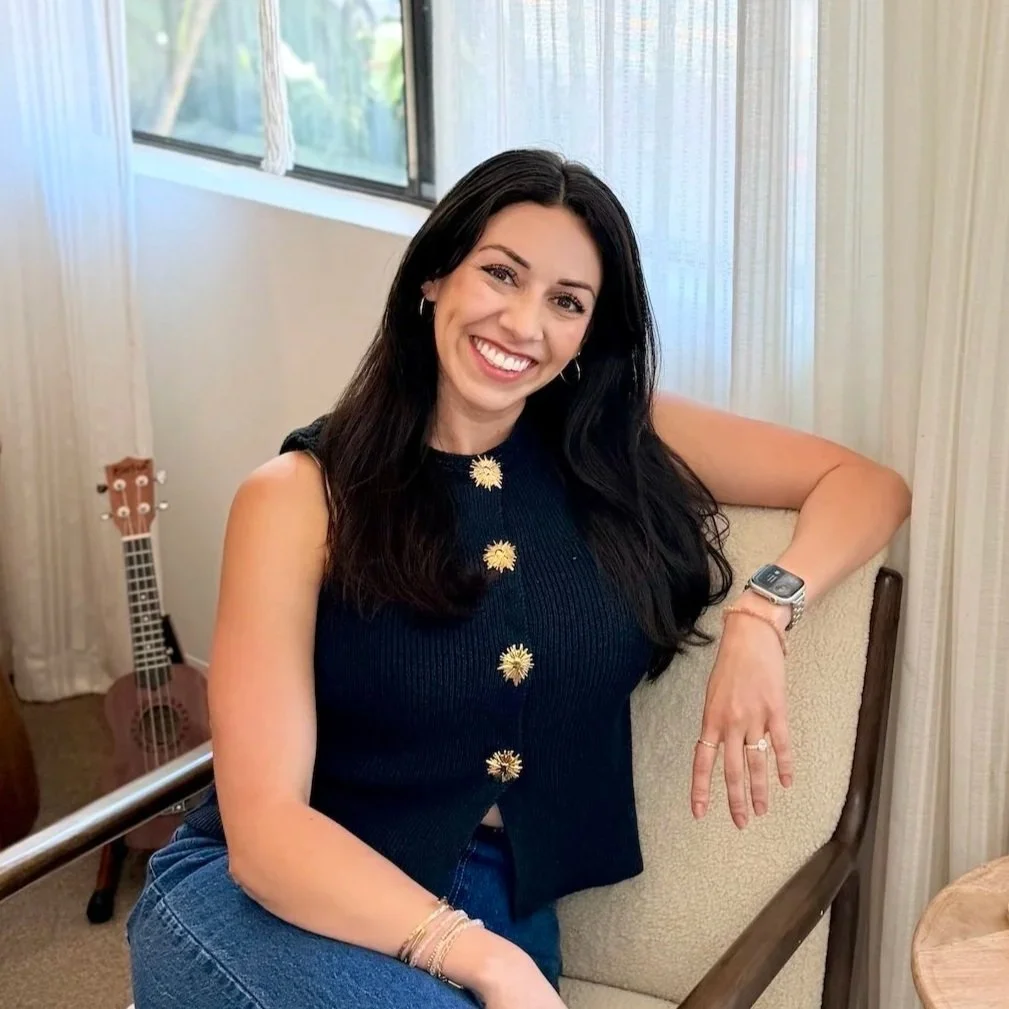 Smiling young woman with long dark hair, wearing a white dress and hoop earrings, sitting outdoors on a grassy area.
