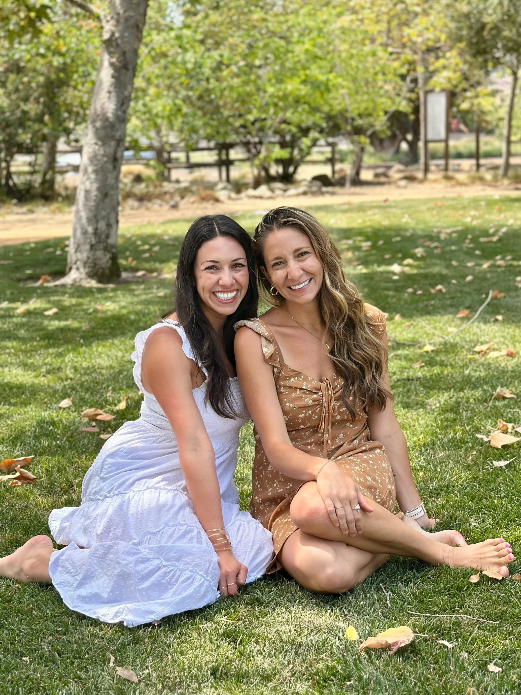 Two women sitting on grass in a park, smiling at the camera, surrounded by trees with fall leaves.