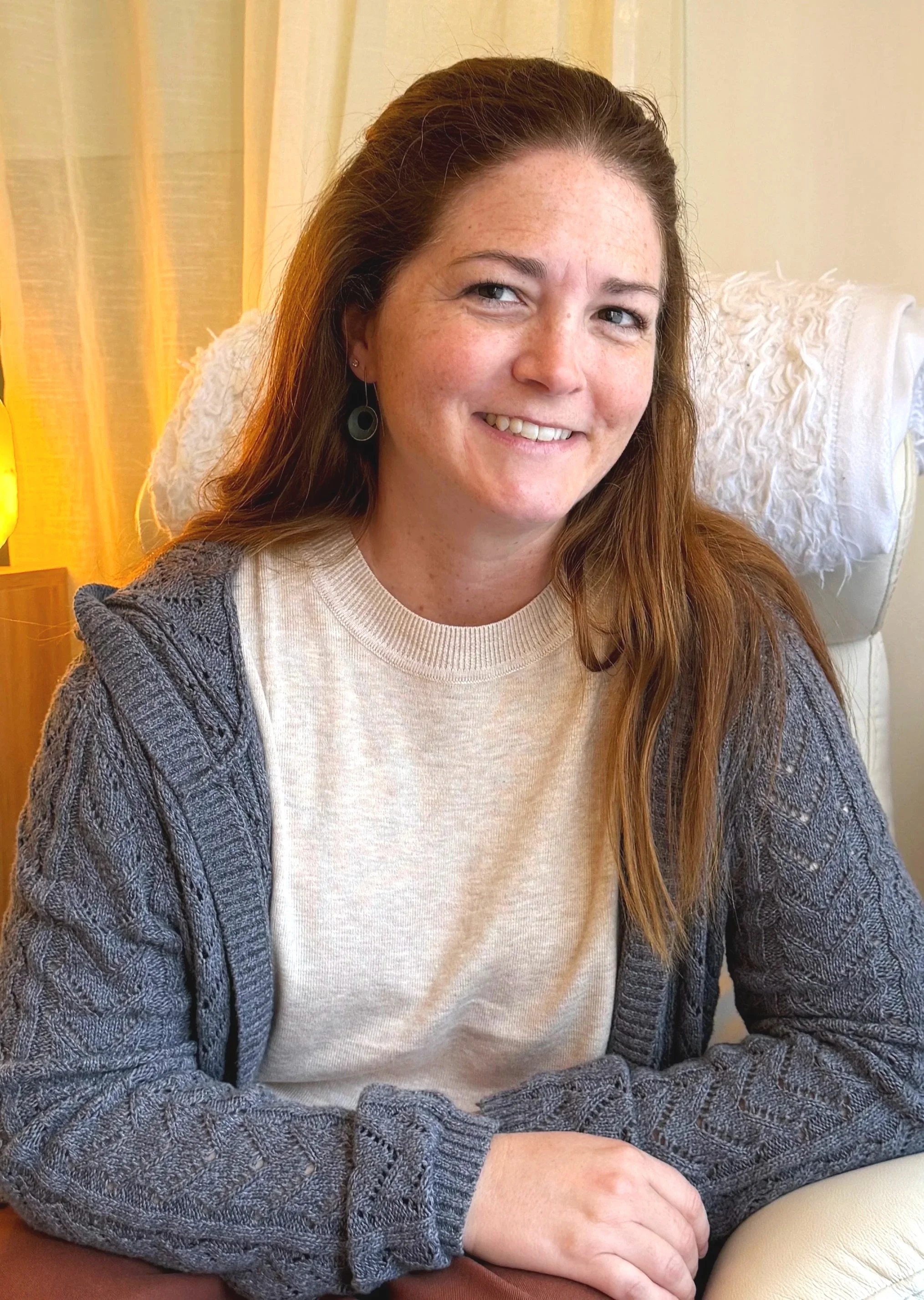 A smiling woman with shoulder-length brown hair, wearing glasses and a cream-colored dress with black and blue spots, sitting in a room with decorative shelves and a yellow patterned box in the background.