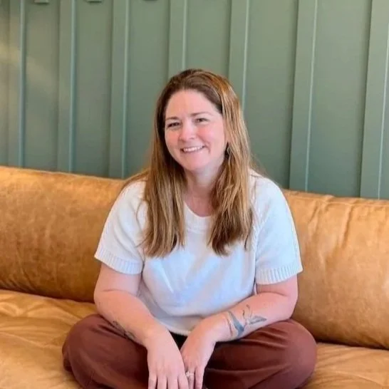 A smiling woman with shoulder-length brown hair, wearing glasses and a cream-colored dress with black and blue spots, sitting in a room with decorative shelves and a yellow patterned box in the background.