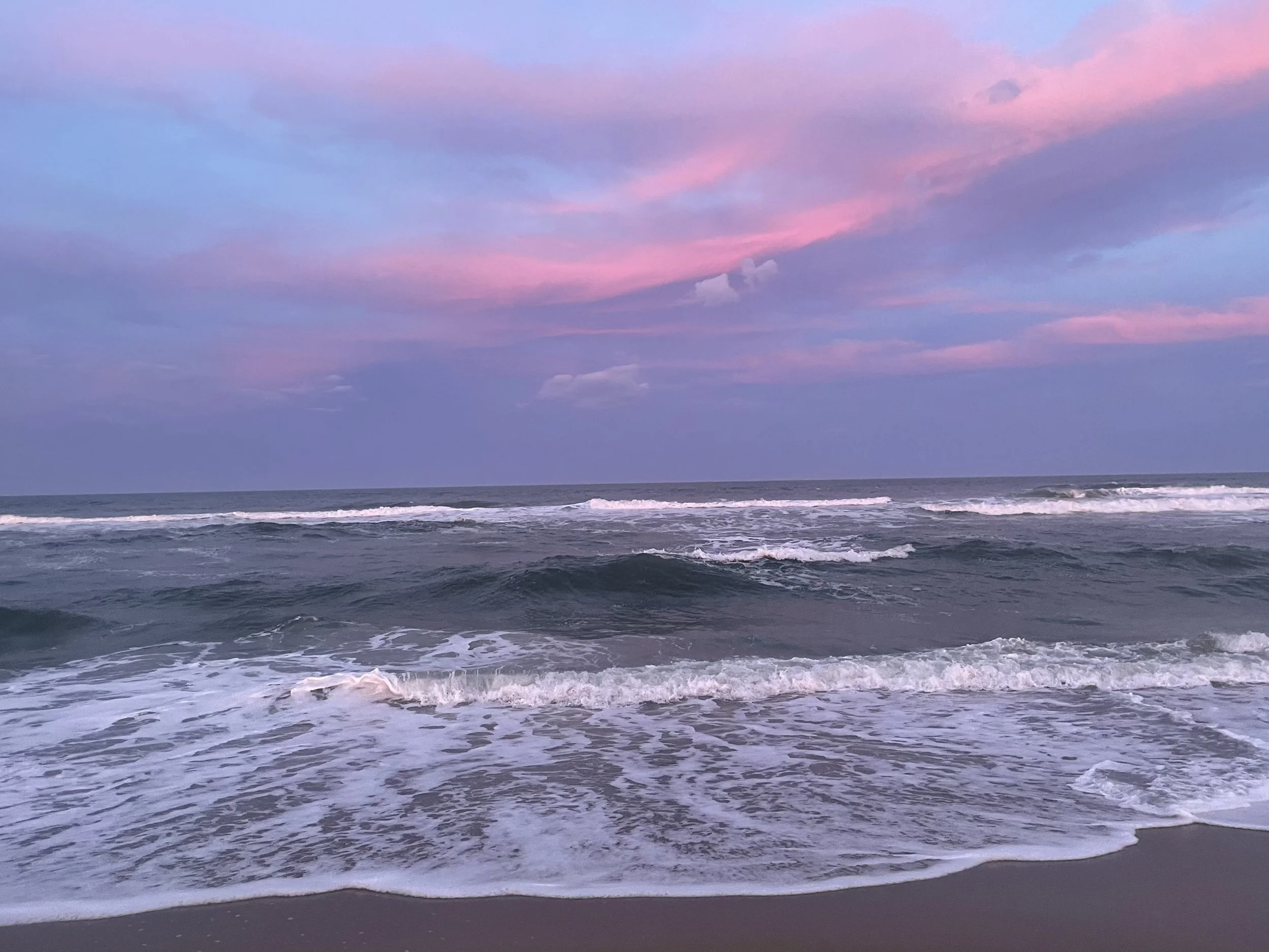 A beach scene during sunset with pink and purple clouds in the sky, gentle waves rolling onto the sandy shore.
