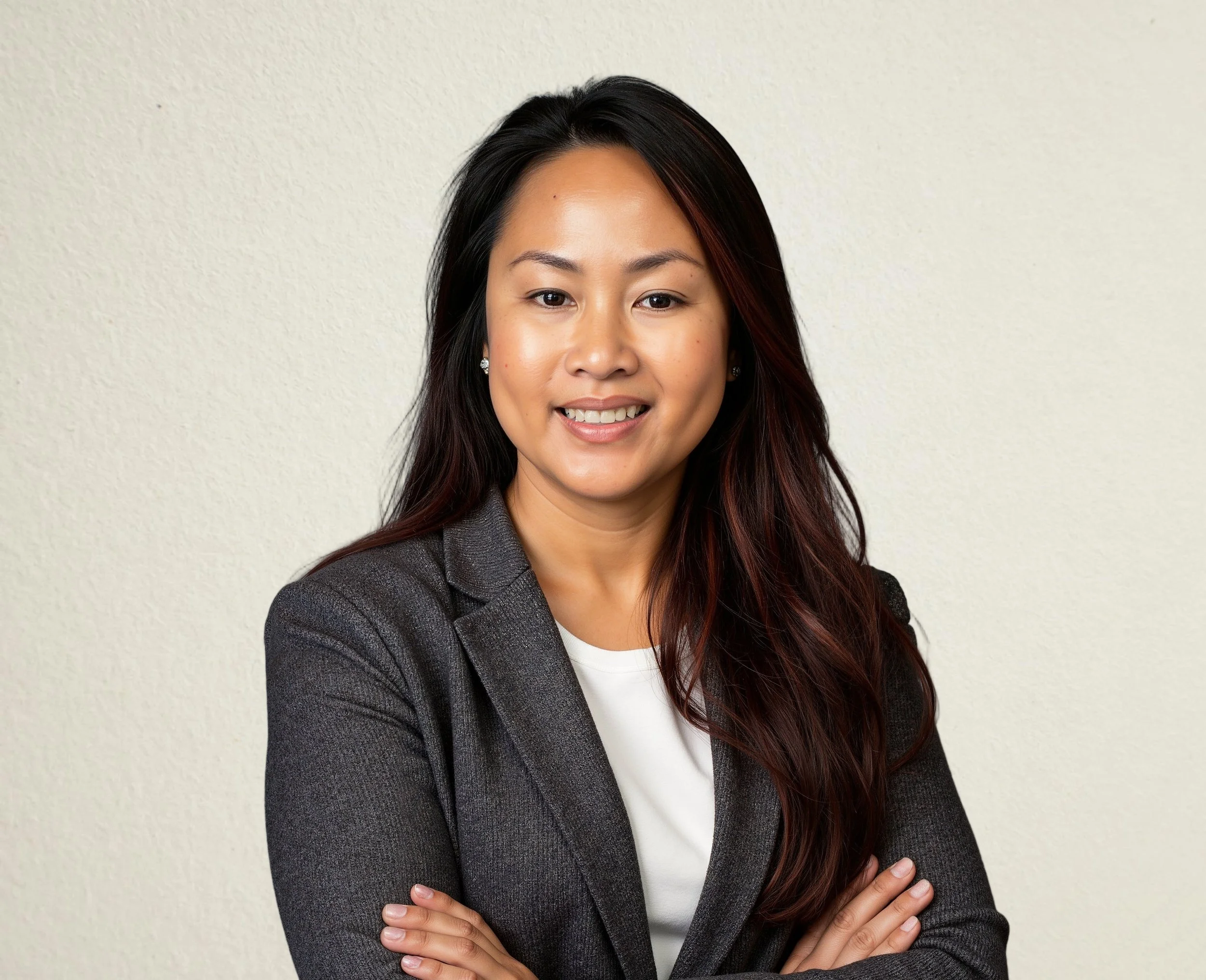 A woman with long dark hair and a friendly smile, wearing a gray blazer over a white shirt standing with crossed arms against a plain beige wall.