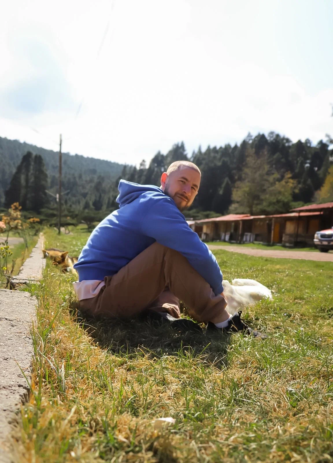 A man sitting on the grass outdoors, wearing a blue hoodie and brown pants, with a dog lying beside him. There are trees and small buildings in the background, with a partly cloudy sky overhead.