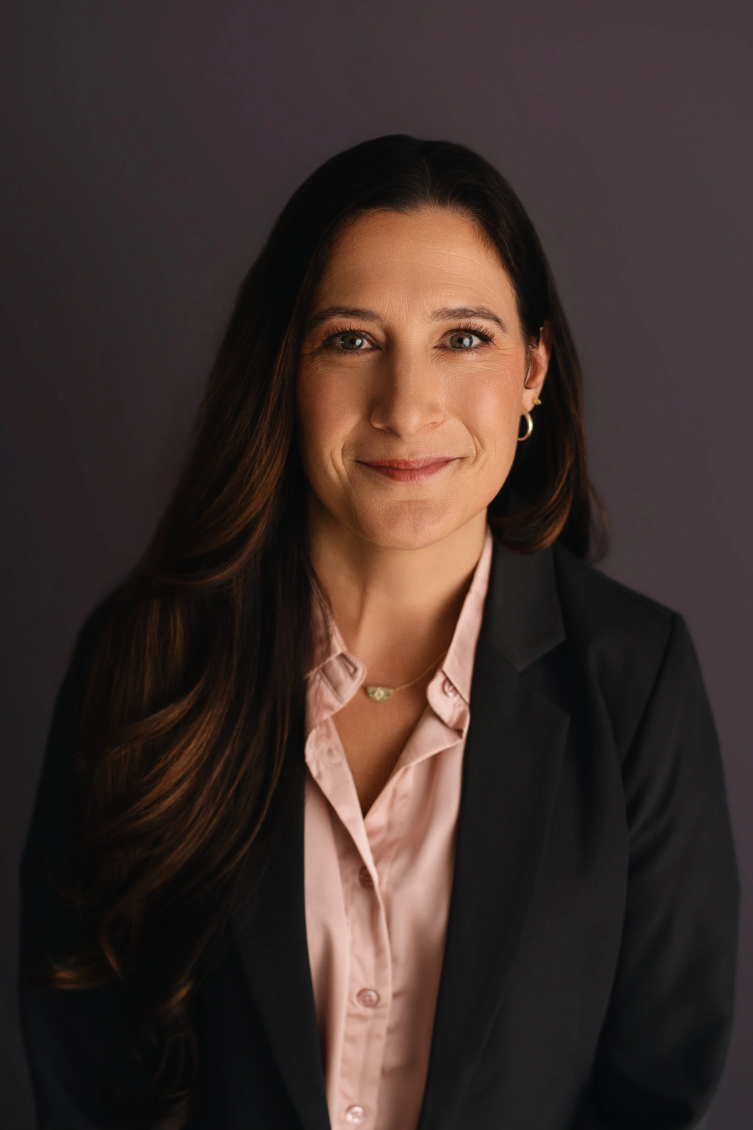 A professional woman with long brown hair and blue eyes, wearing a black blazer and a light pink blouse, smiling against a dark gray background.