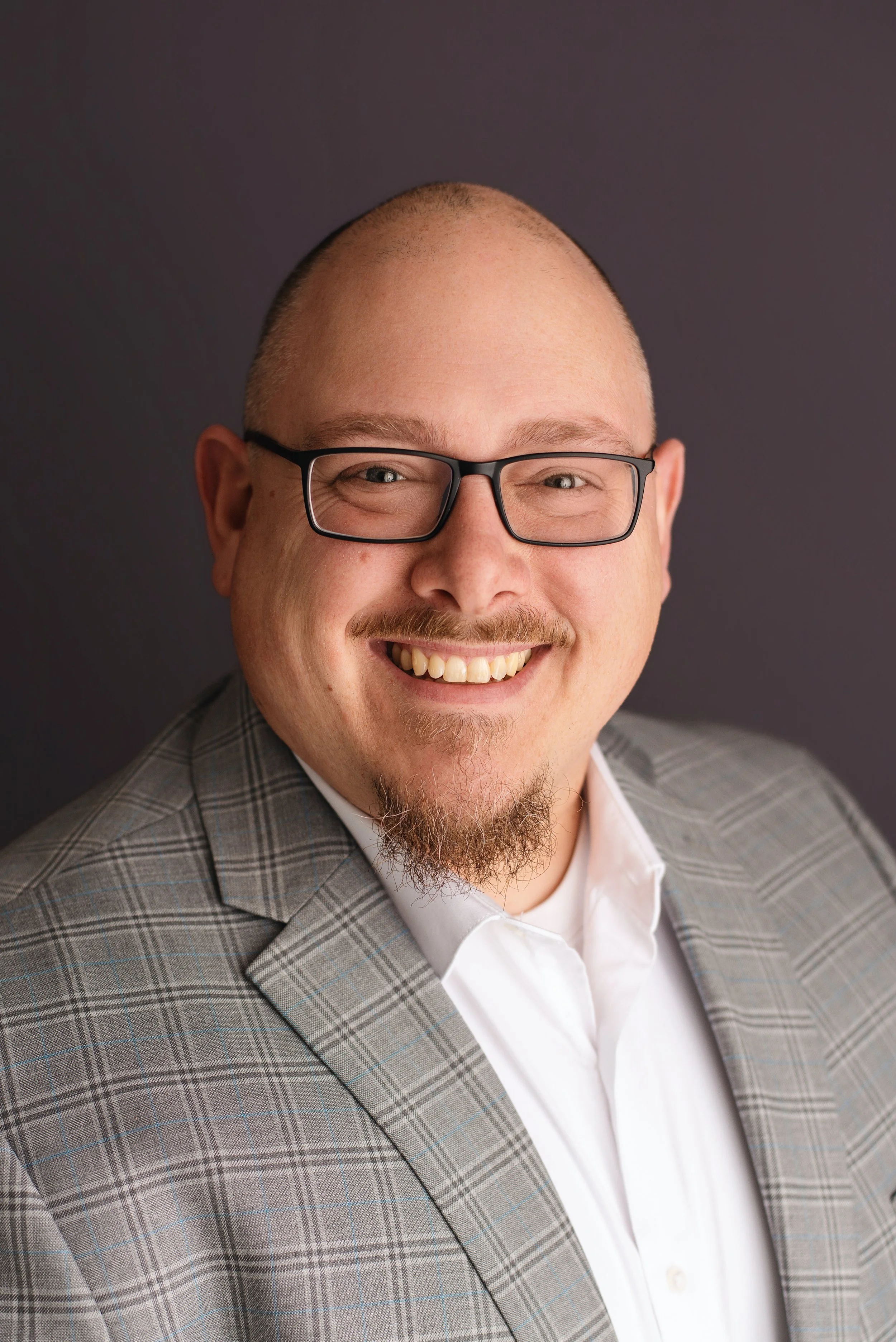 Close-up of a smiling man with glasses, a checkered blazer, and a white shirt against a dark background.