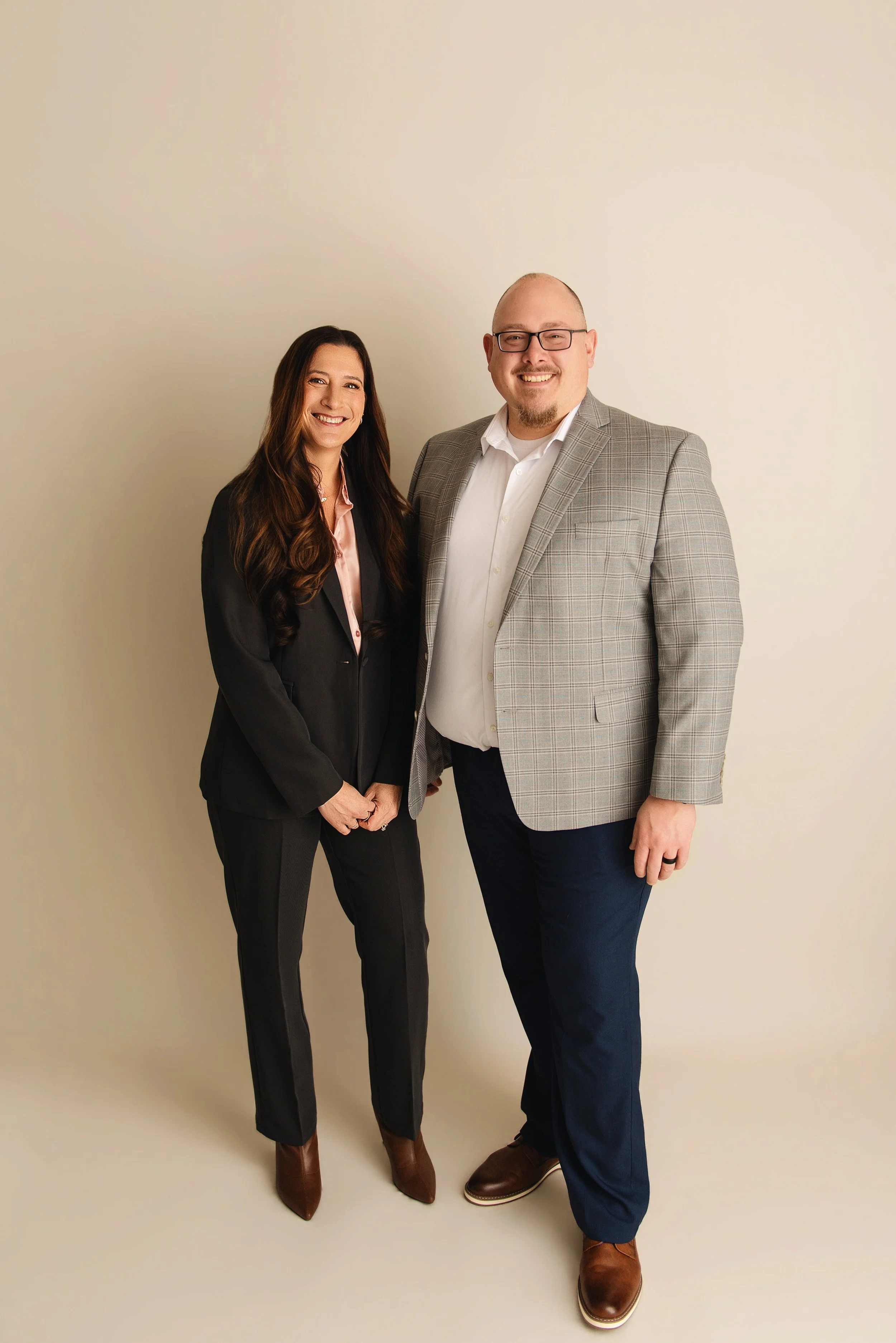 A woman with long brown hair wearing a black suit and brown boots standing next to a man with glasses, a beard, wearing a gray checked blazer, white shirt, dark blue pants, and brown shoes, smiling together against a plain beige wall.