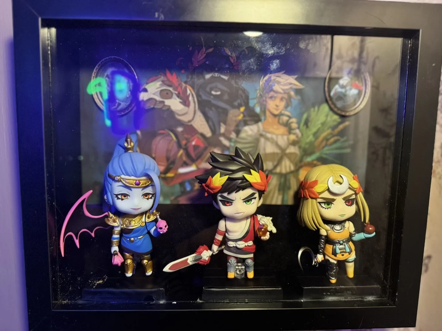 Hades Diorame featuring Zagreus, Megera and Melinoe Nendroids in a display case, with a colorful illustrated background showing Hades, Persophone inside a black frame.
