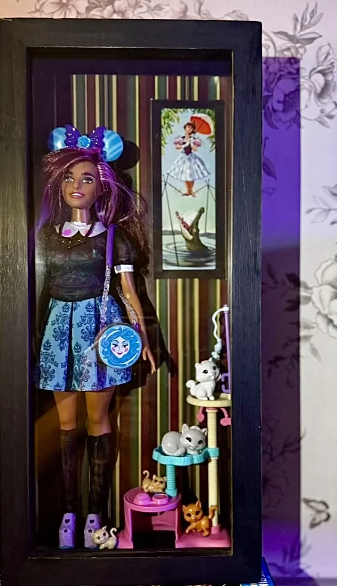 Display case containing a doll with long brown hair, a black and blue dress, knee-high socks, and a blue Mickey ears. The background is the Stretching Room of the Haunted Mansion. There is a Painting of the tightrope walker. There are small animal fi