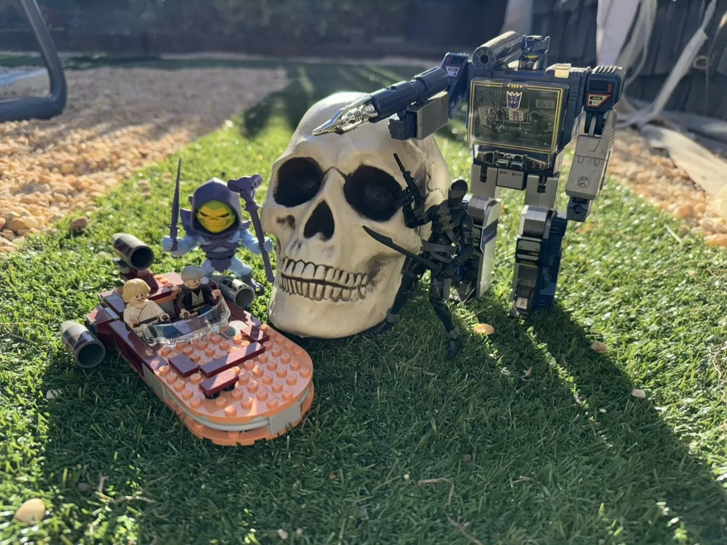 A collection of toys including a LEGO Star Wars speeder bike with two minifigures, a large skull, and a robot with Decepticon symbols, arranged outdoors on grass with sunlight.