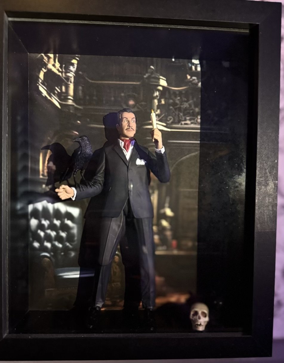 A figure of Vincent Price n a tuxedo holding a candle and a black crow standing on his arm inside a display case. There is a skull on the floor of the case and a dark, ornate backdrop.