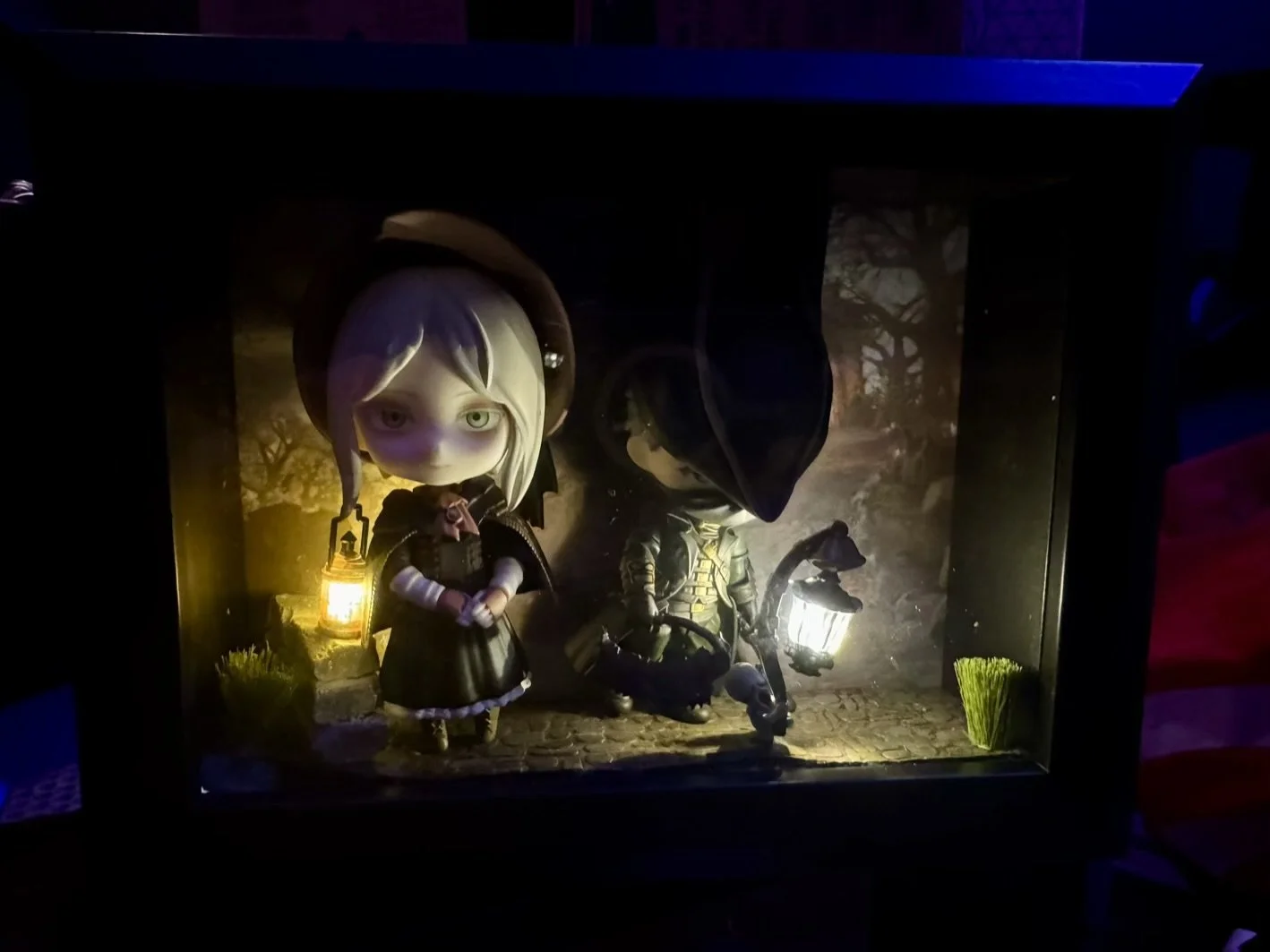 Miniature diorama scene of Bloodborne with two Nendoroids includeing the Dolls, the Hunter, lanterns, and a background of trees at dusk, inside a glass display case.