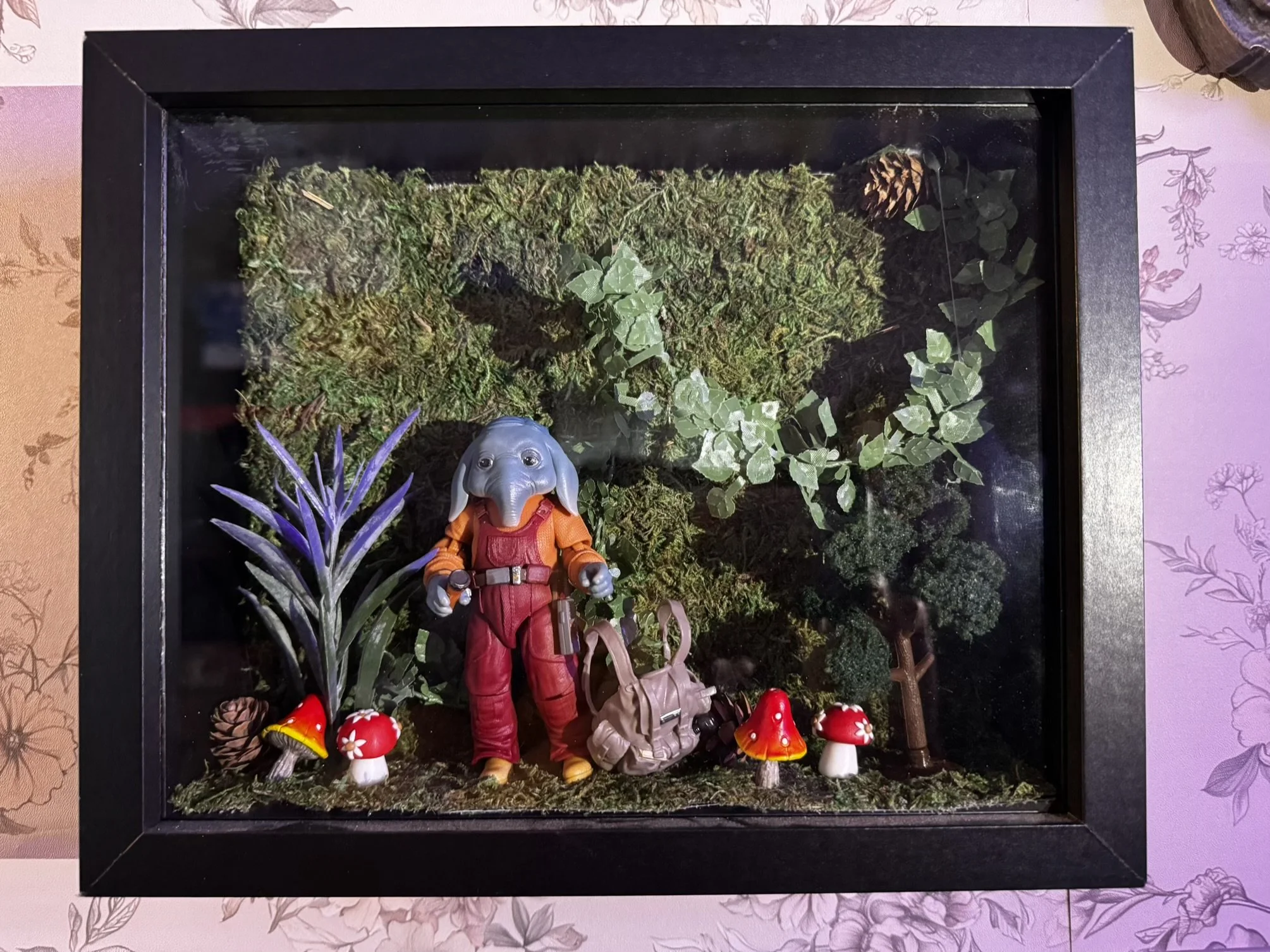 A diorama of Star Wars Skeleton Crew with a Neil figure in red overalls, surrounded by plants, mushrooms, moss, and pinecones, enclosed in a black frame.