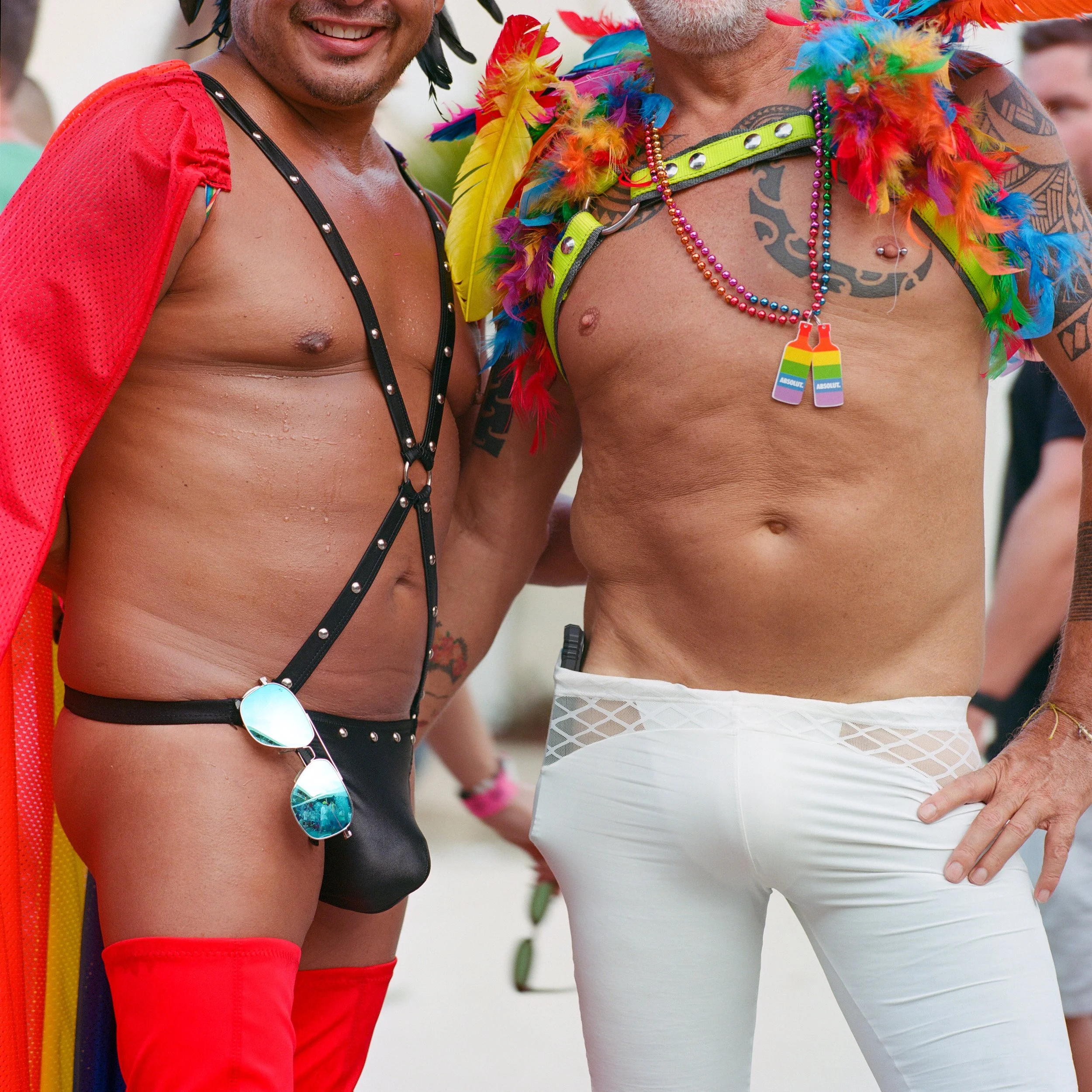 Two men dressed in colorful, flamboyant outfits celebrating at an event. One wears a red cape and a black harness with sunglasses hanging from it, while the other is shirtless with rainbow feathered accessories and white pants.