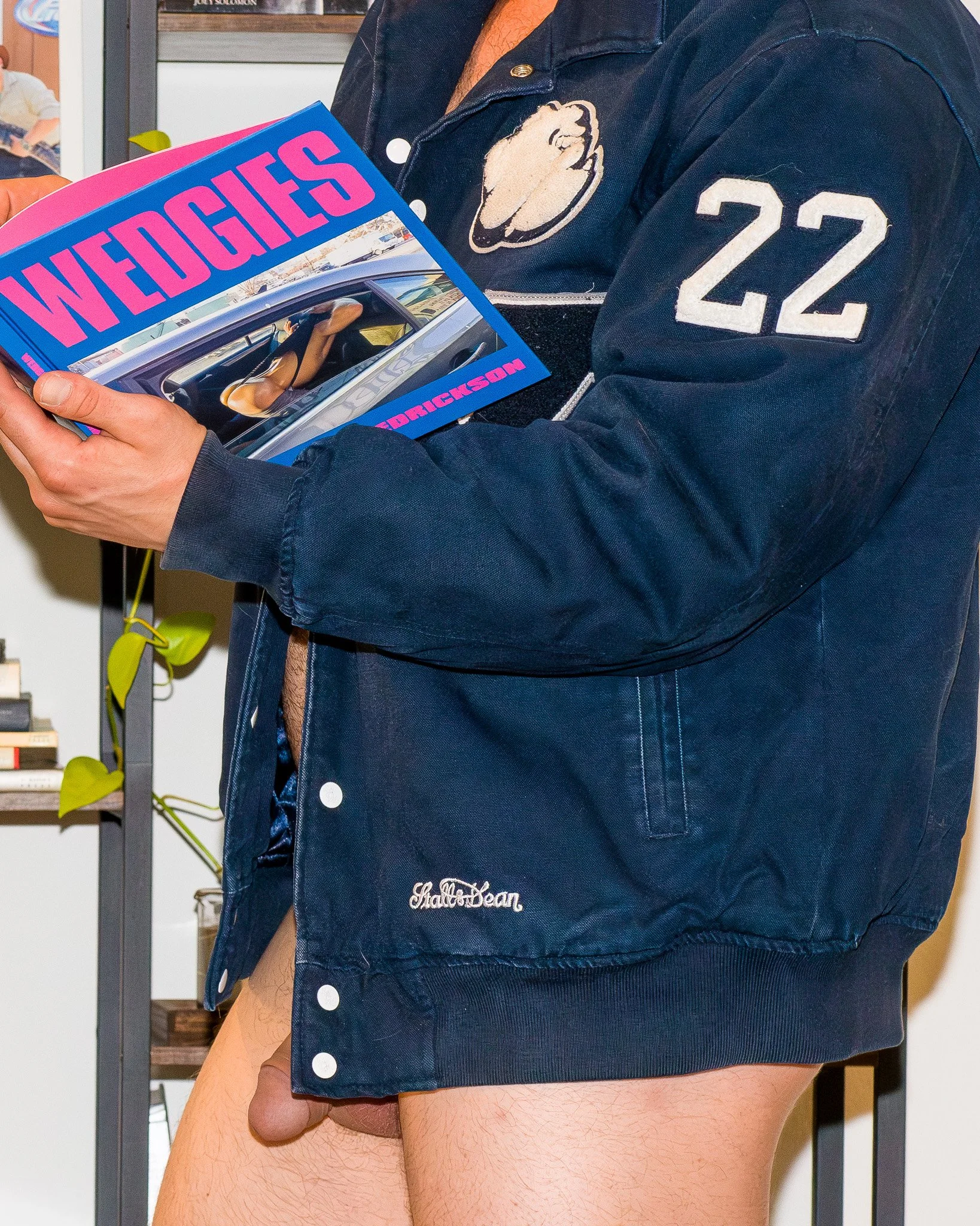 A person wearing a blue letterman jacket holding a magazine titled 'WEDGIES' with a photo of a woman wearing black shoes, with the person's pants pulled down, exposing their genitals.