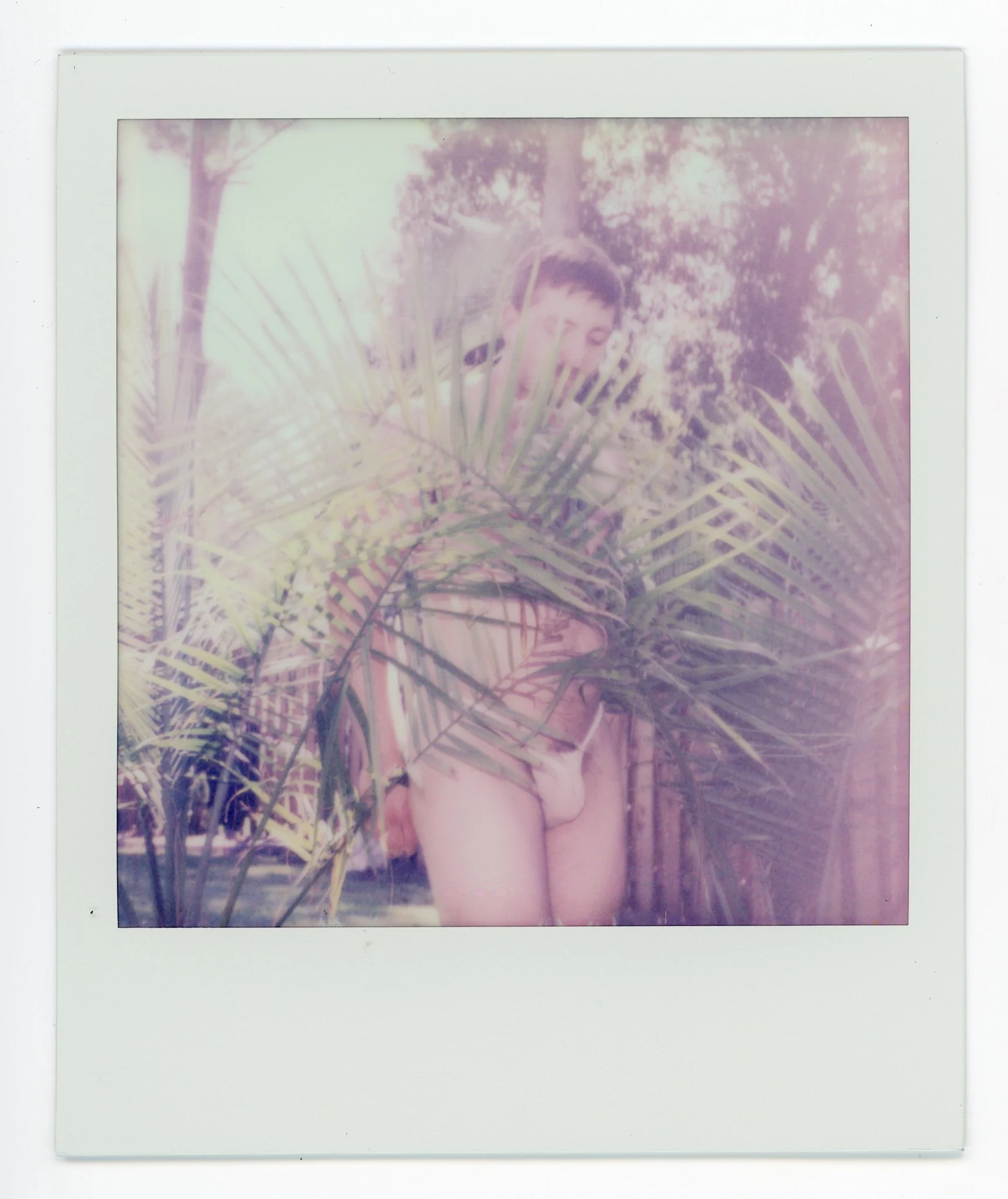 A Polaroid photograph of a young person standing behind large tropical leaves outdoors, with trees and sky in the background, in soft, faded colors.