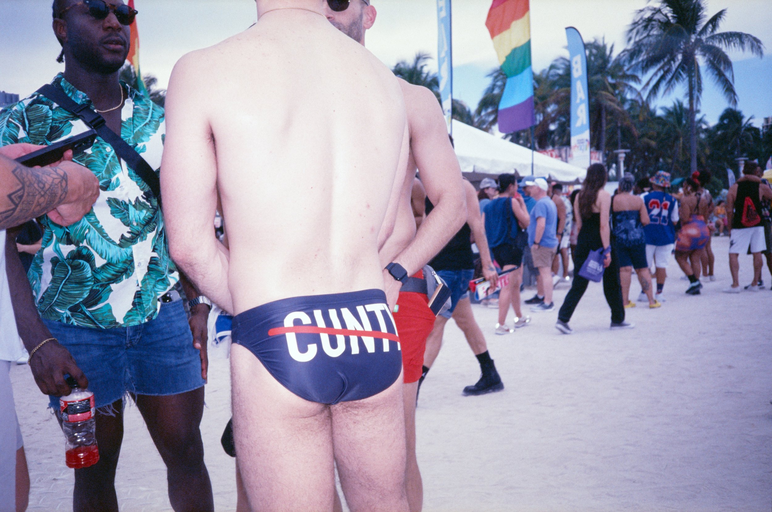 Person standing on the beach wearing underwear with large text, surrounded by crowds at a festival with palm trees, tents, and rainbow flags in the background.