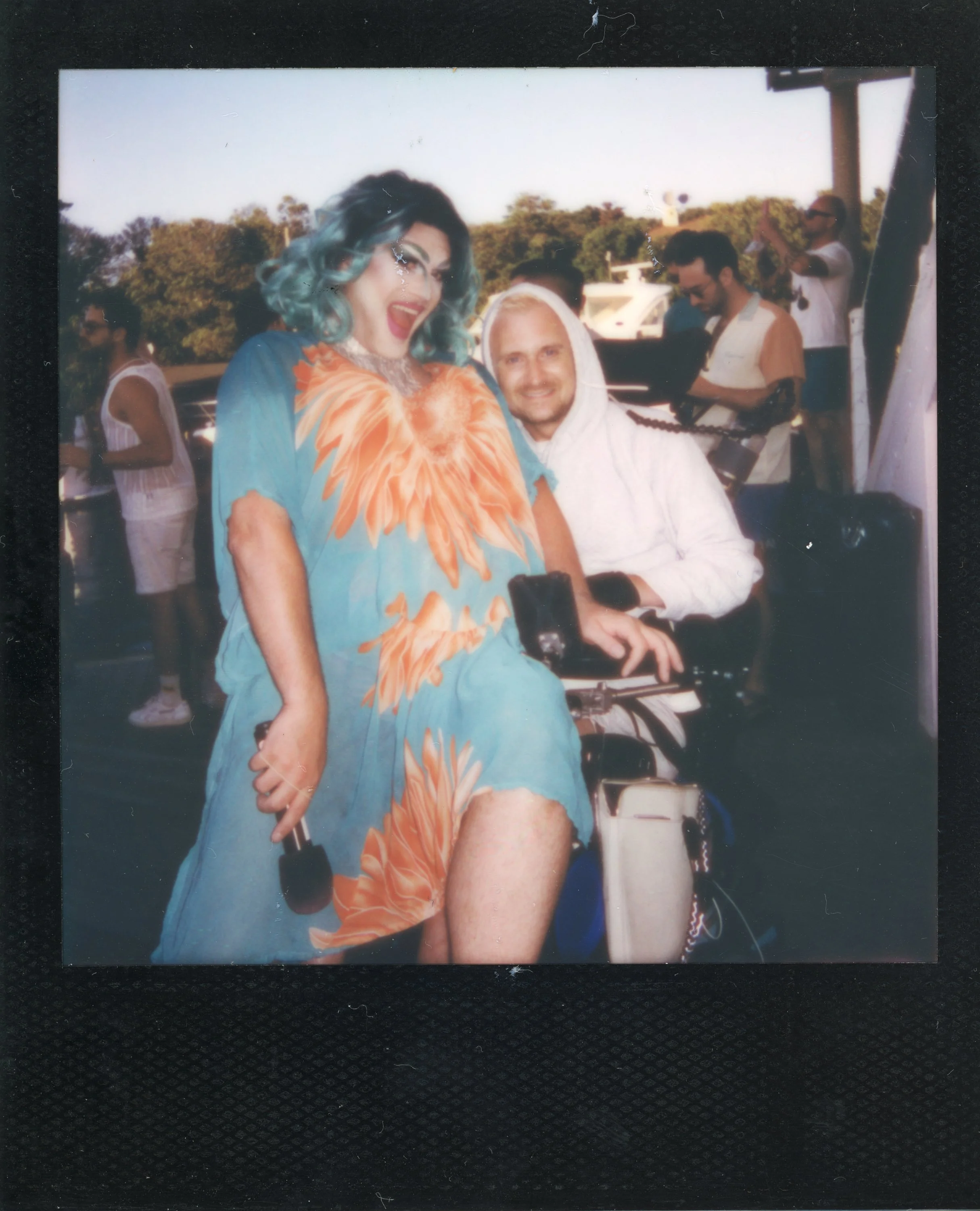 Person with teal hair in a colorful dress filling the frame, smiling and holding a microphone, with a person in a white hoodie sitting on a bicycle behind them. Several other people are in the background outdoors, some taking photos.