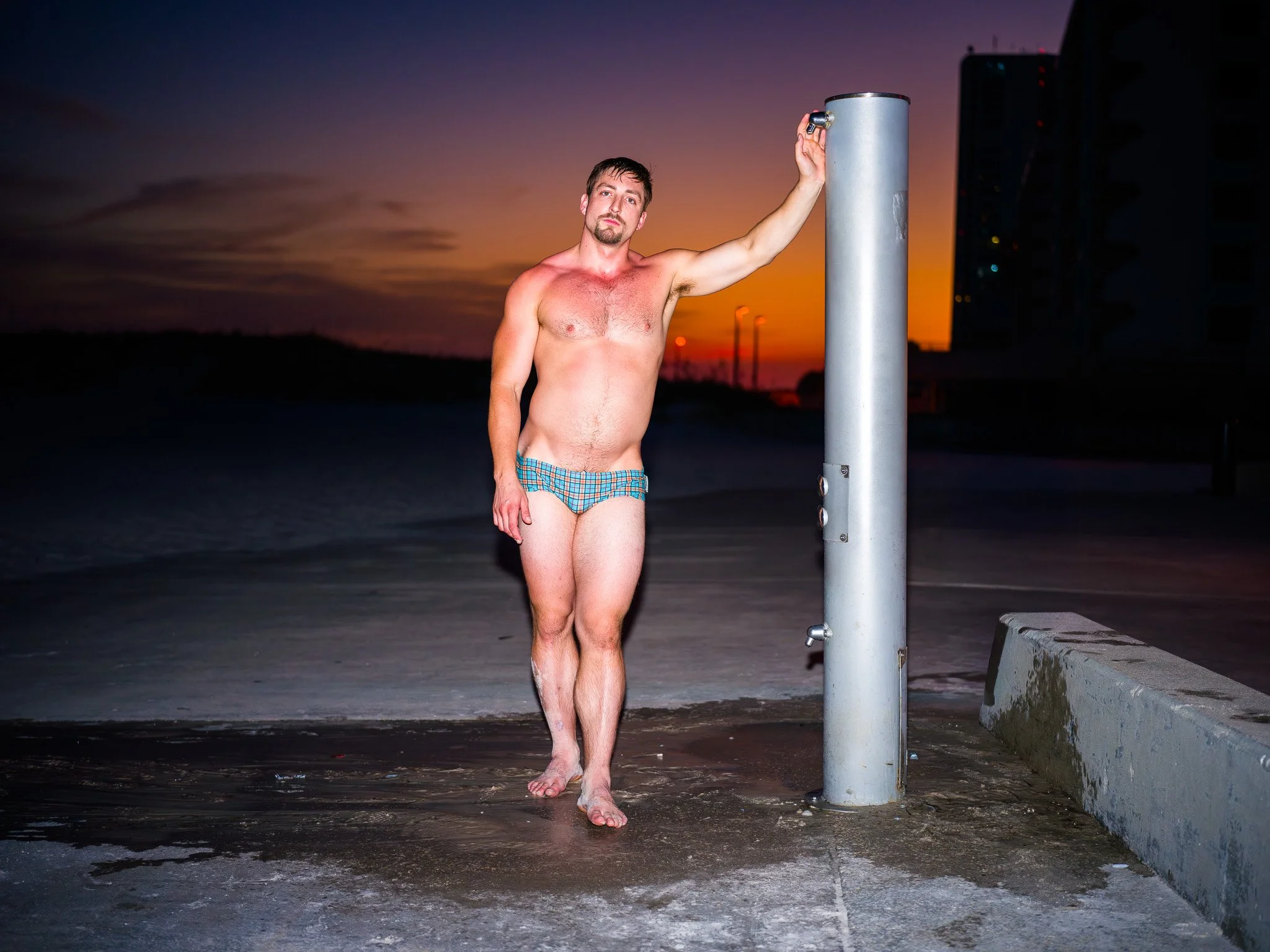 A shirtless man wearing swim trunks standing barefoot at an outdoor shower on a beach during sunset.