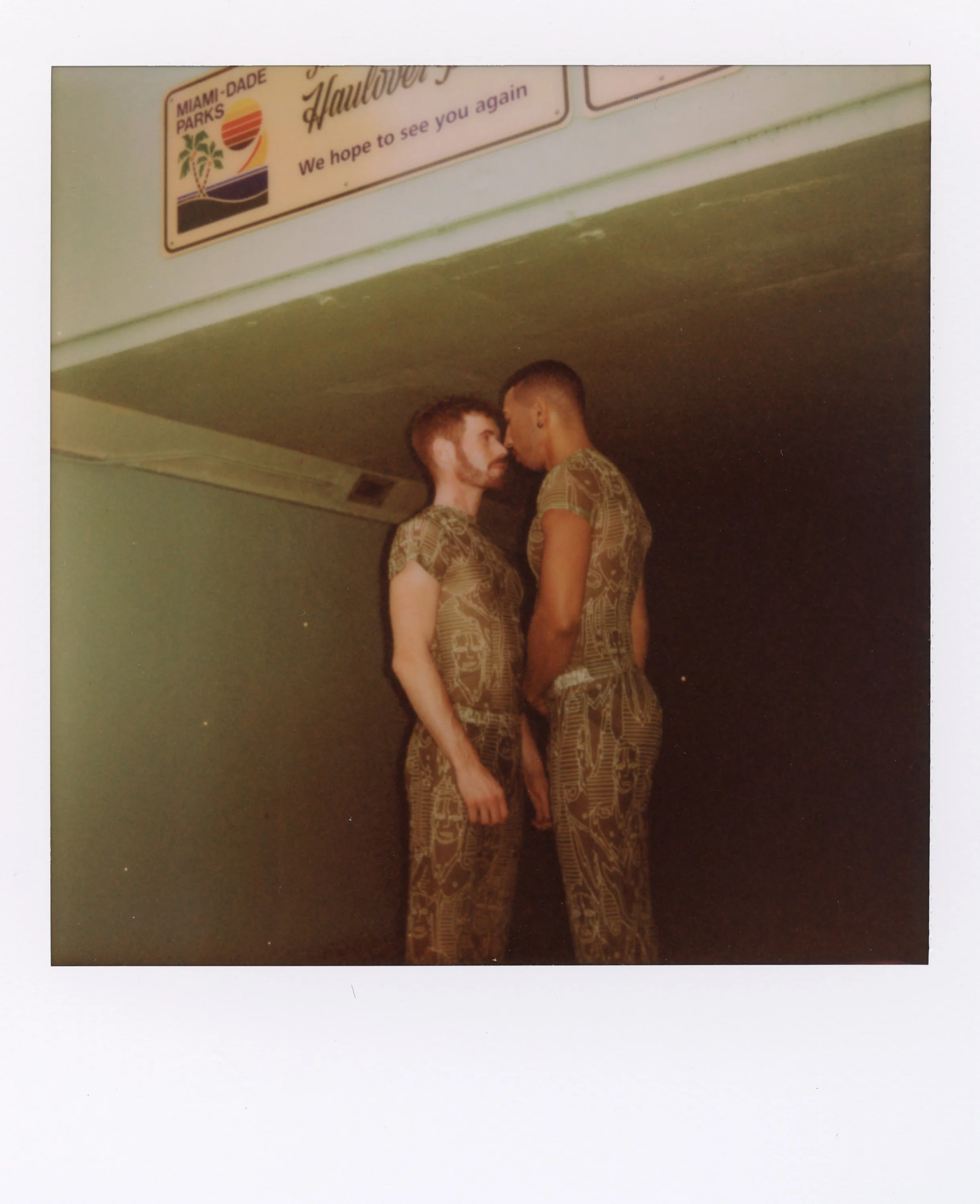 Two women wearing matching patterned outfits standing very close with foreheads touching in a dimly lit room.