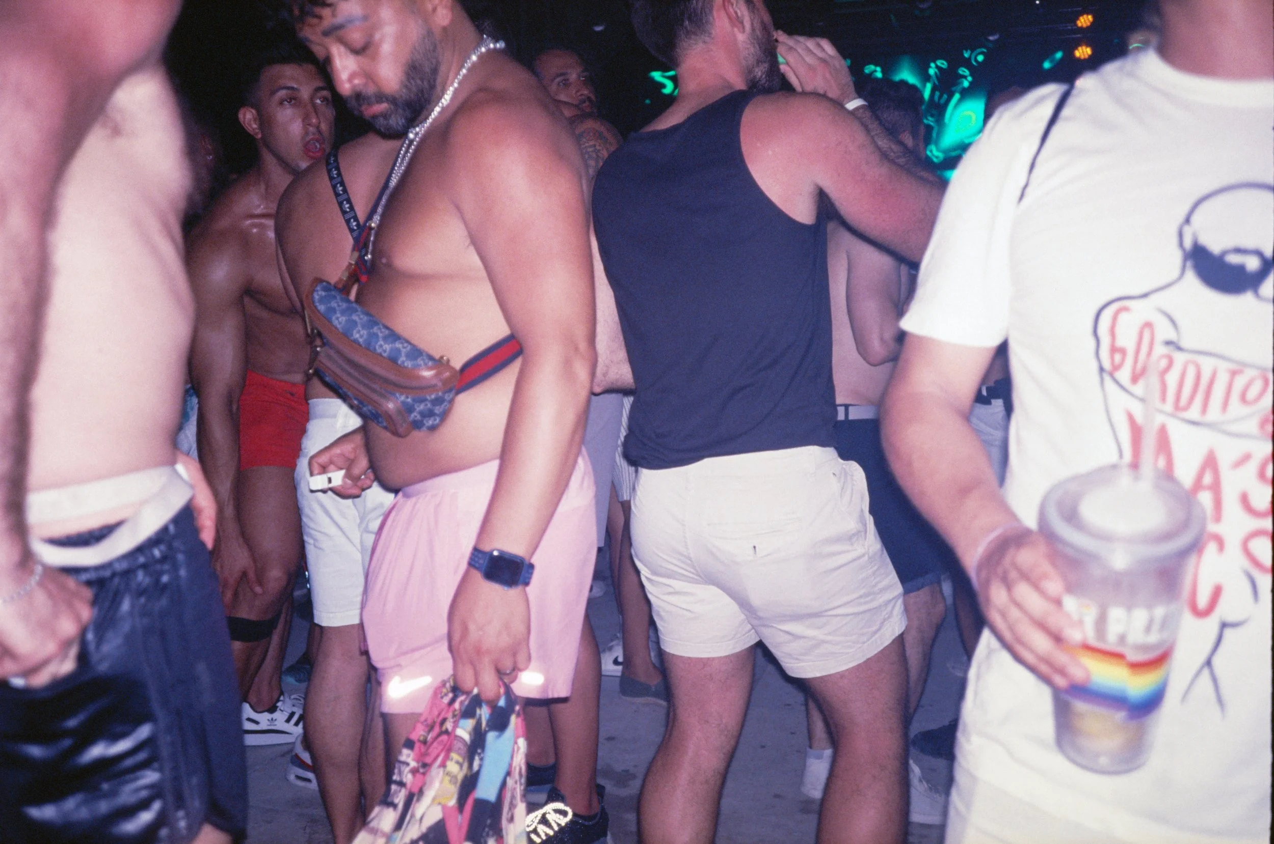 People dancing and socializing at a party or nightclub, casual summer attire, some shirtless, colorful lighting.
