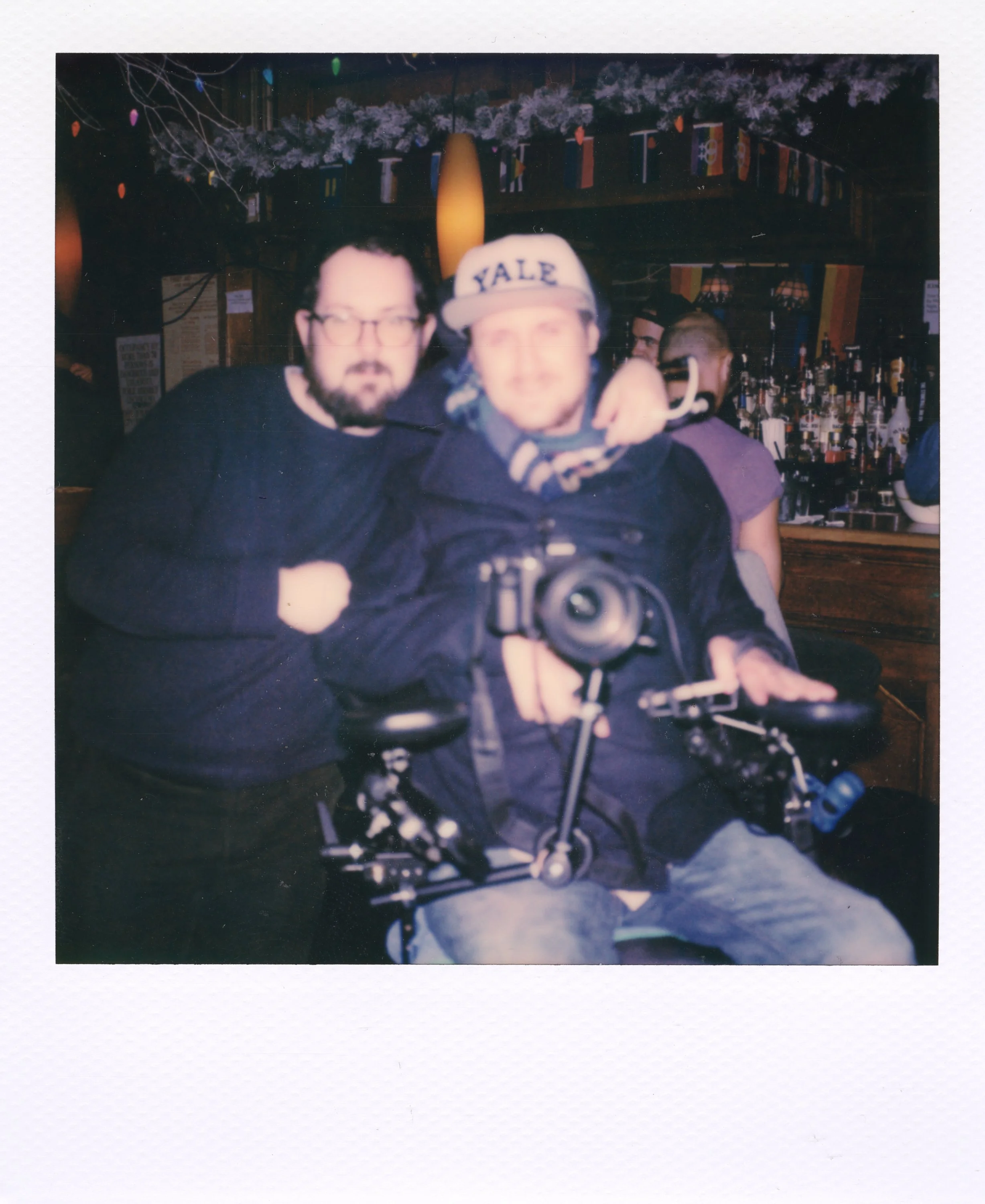 Two men sitting at a bar with one behind the wheelchair, both posing for a photo. The man in front is holding a camera and is wearing a Yale cap. The background features bar shelves with bottles and national flags on a string.