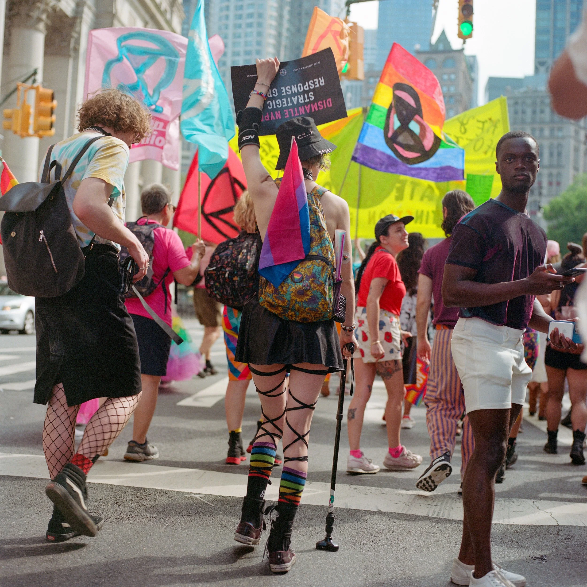 Crowd of diverse people participating in a pride parade, holding colorful flags and signs against urban city backdrop.