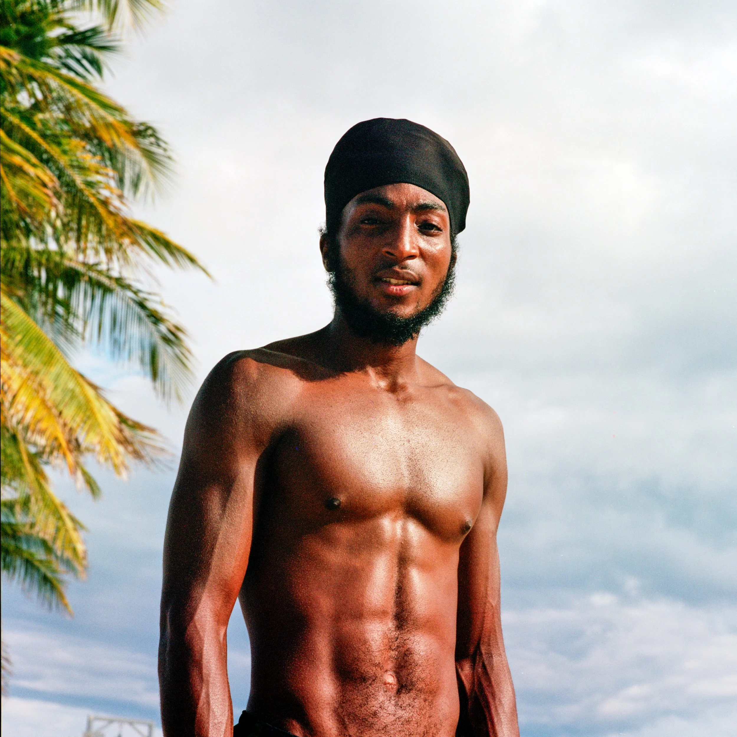 A shirtless man with a beard and a black headwrap standing outdoors near palm trees with a cloudy sky in the background.
