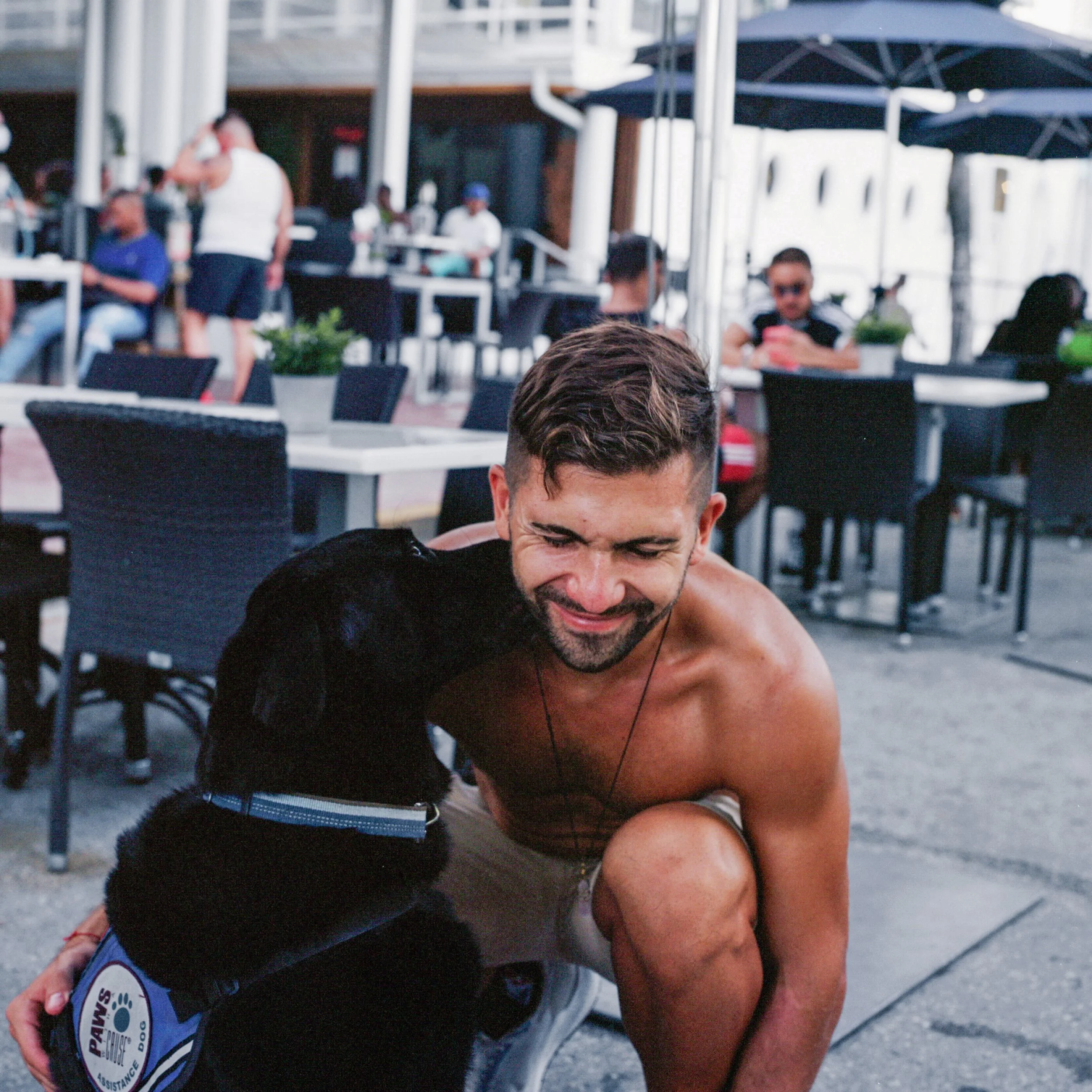 A shirtless man crouching down and smiling as a black dog wearing a service dog vest affectionately sniffs his face at an outdoor café with tables, chairs, and other patrons in the background.