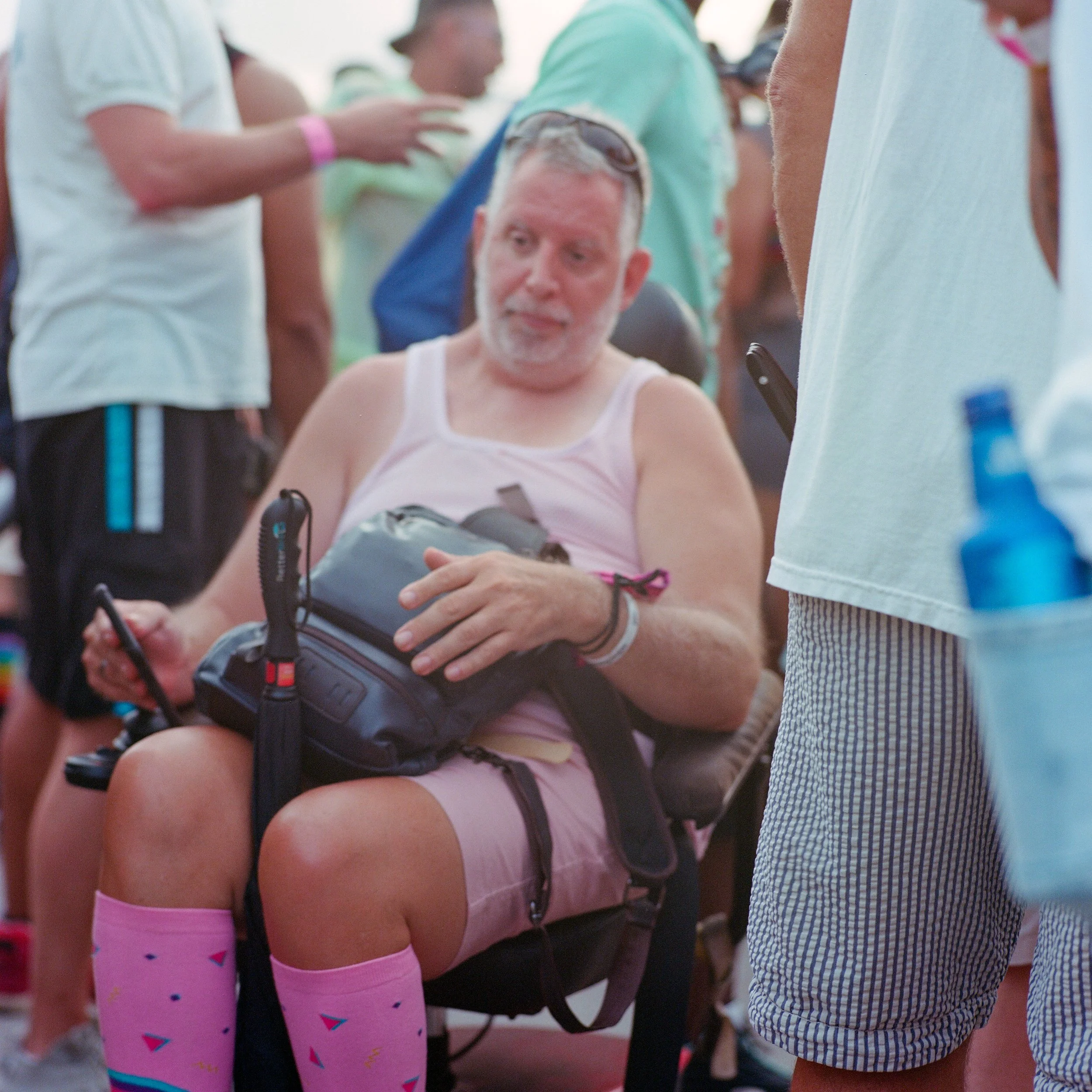 A man sitting in a chair at an outdoor event, wearing pink socks and shorts, holding a smartphone and a black bag, surrounded by people in casual clothing.
