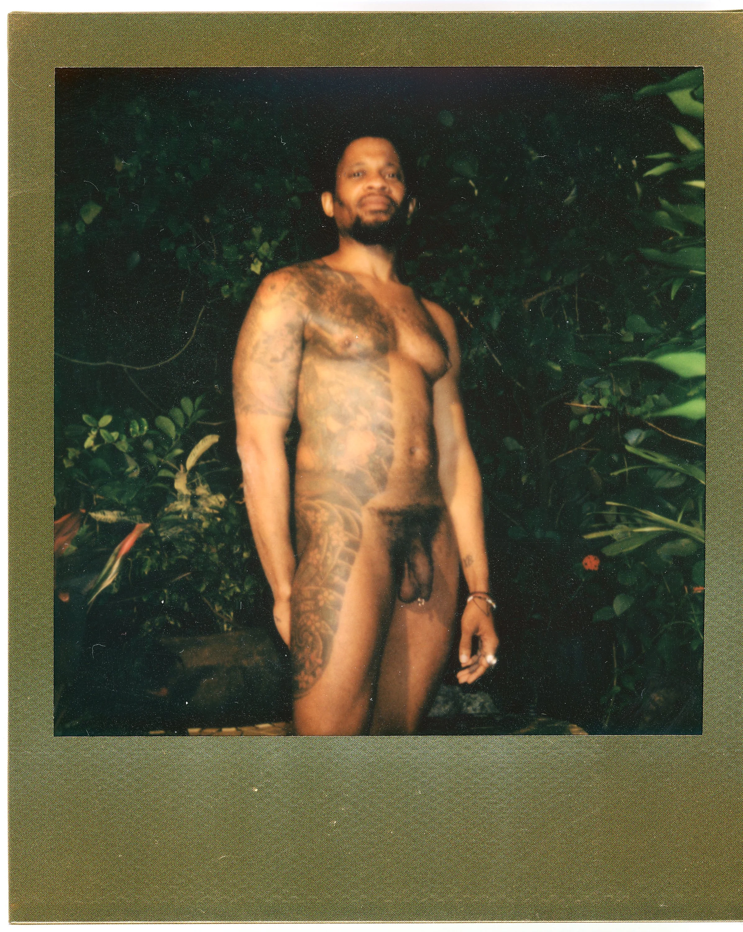A man with tattoos standing outdoors against green foliage, nude except for tattoos, jewelry, and rings.