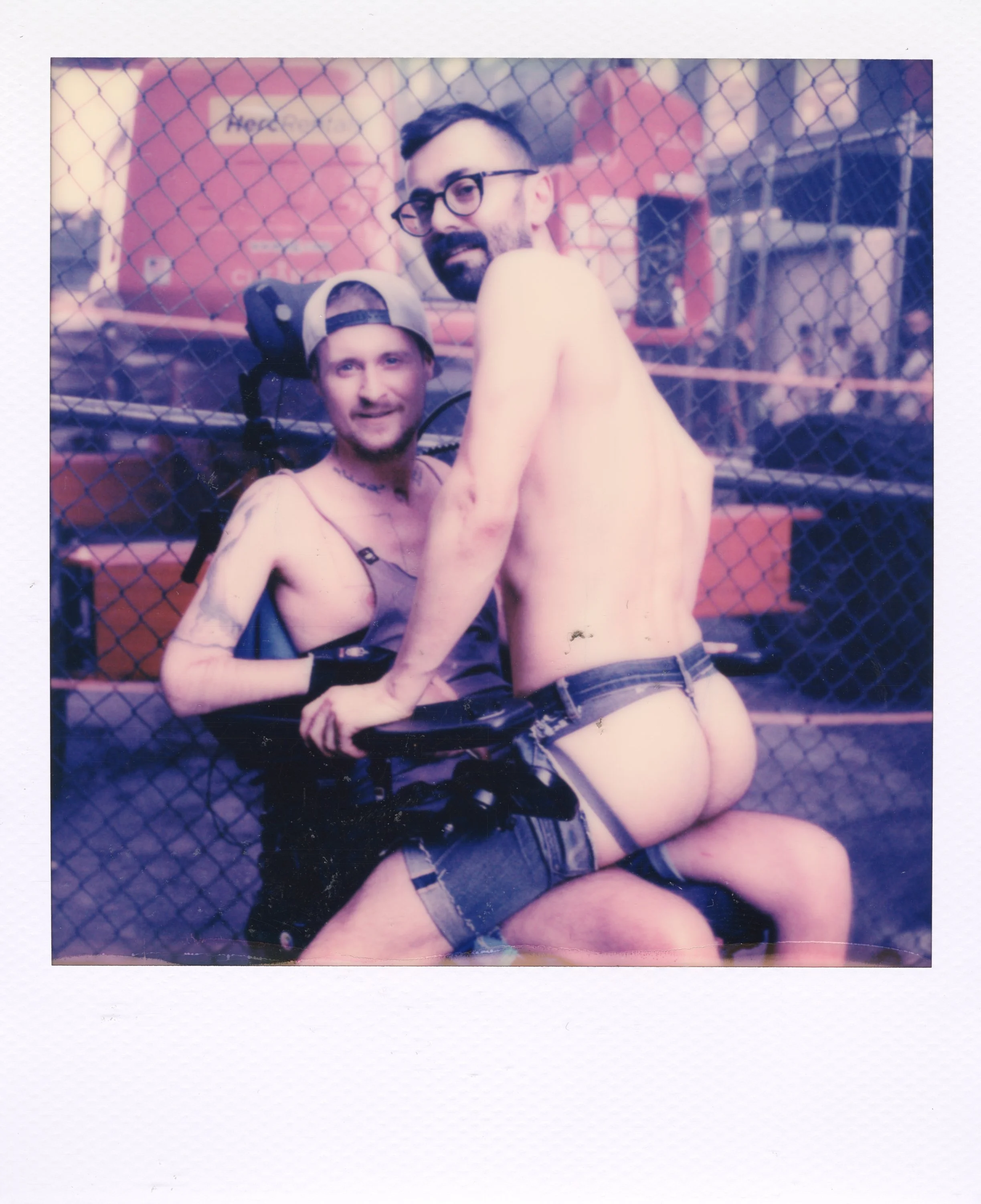 Two men posing behind a chain-link fence, one sitting in a wheelchair and the other naked from the waist down, with a background of orange equipment and a structure.