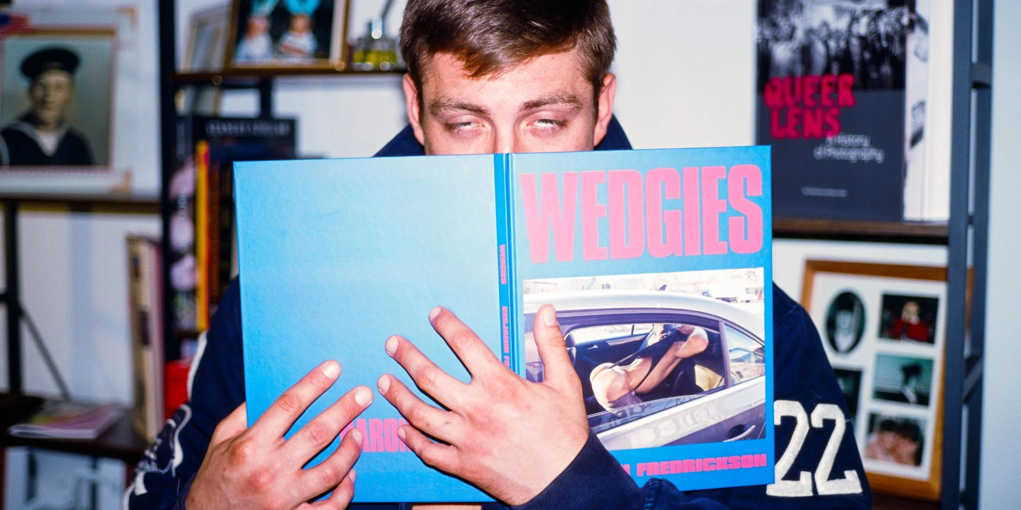 A person with light brown hair reads a large blue book titled 'WEDGES' in a room with shelves of framed photos and books, some titled 'Queer Lens'.