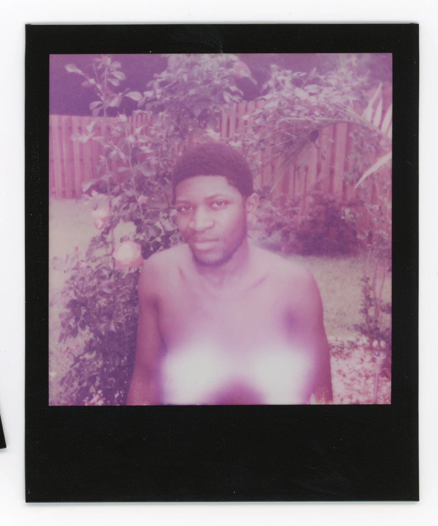 A young man with dark skin and short curly hair, shirtless, standing outdoors in a garden with plants and a wooden fence, looking at the camera.