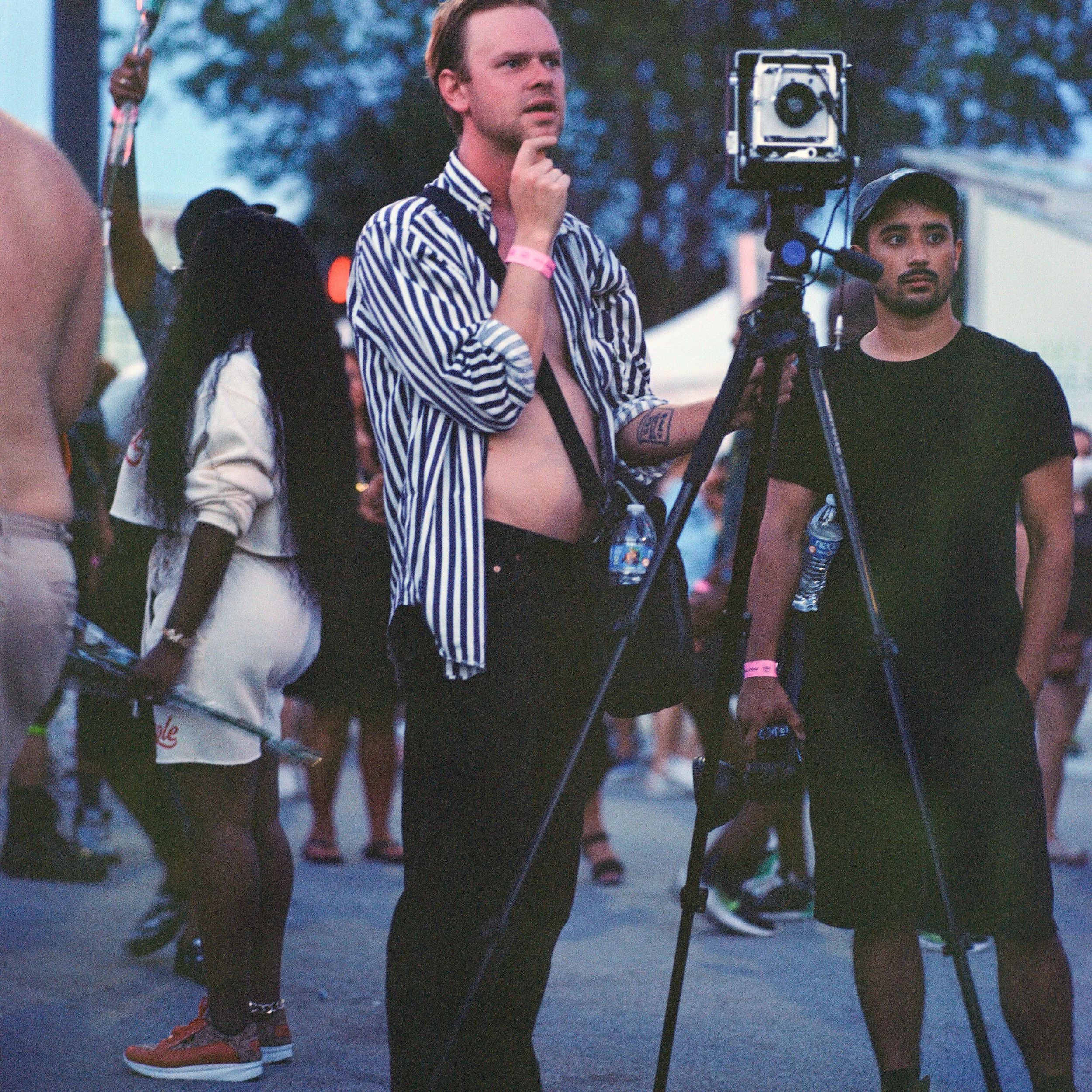 Two men operating a camera at an outdoor event during dusk, surrounded by people, with tents and trees in the background.