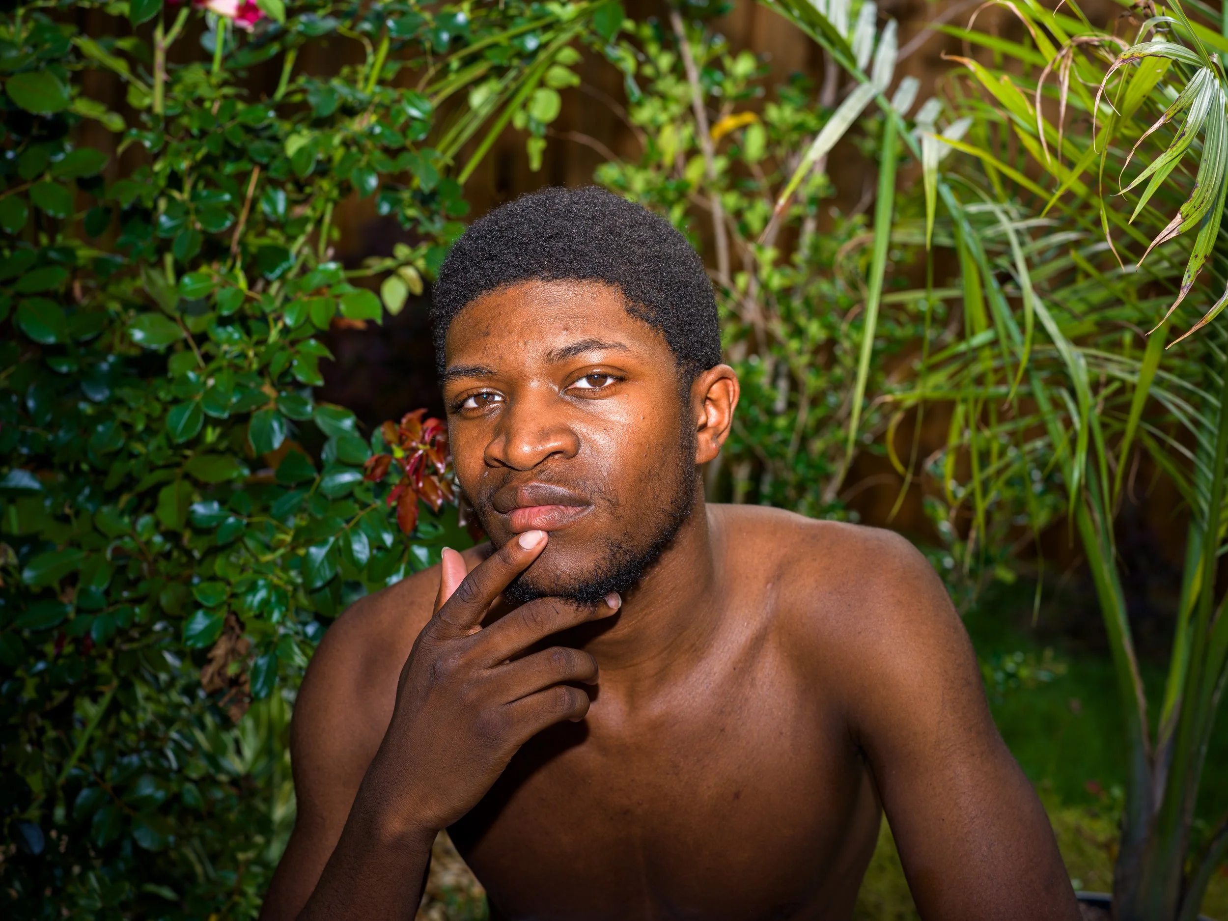 A shirtless young man with short curly hair and a beard poses confidently in front of lush green foliage, resting his chin on his hand.