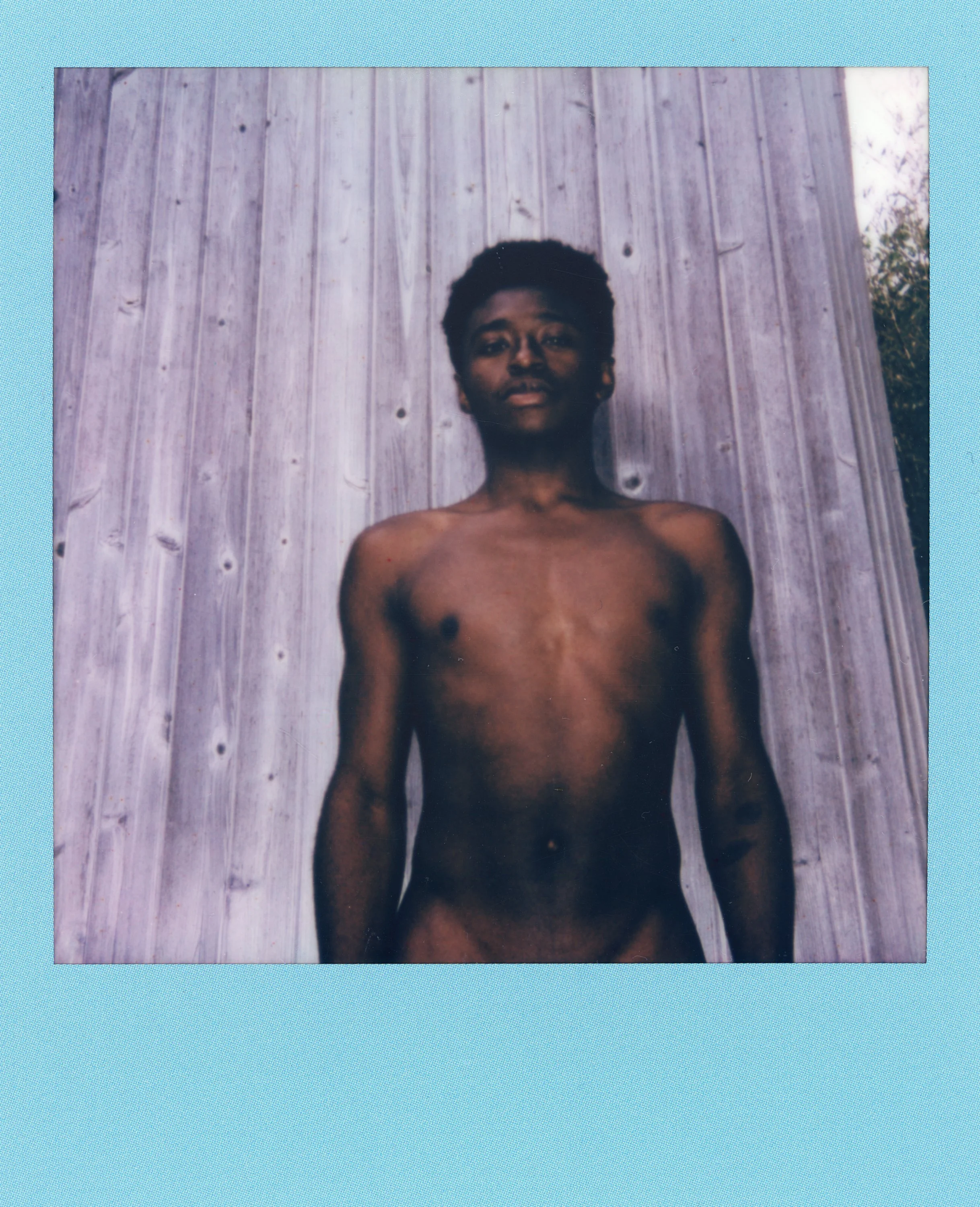 A young person standing shirtless in front of a wooden background, with a neutral expression.