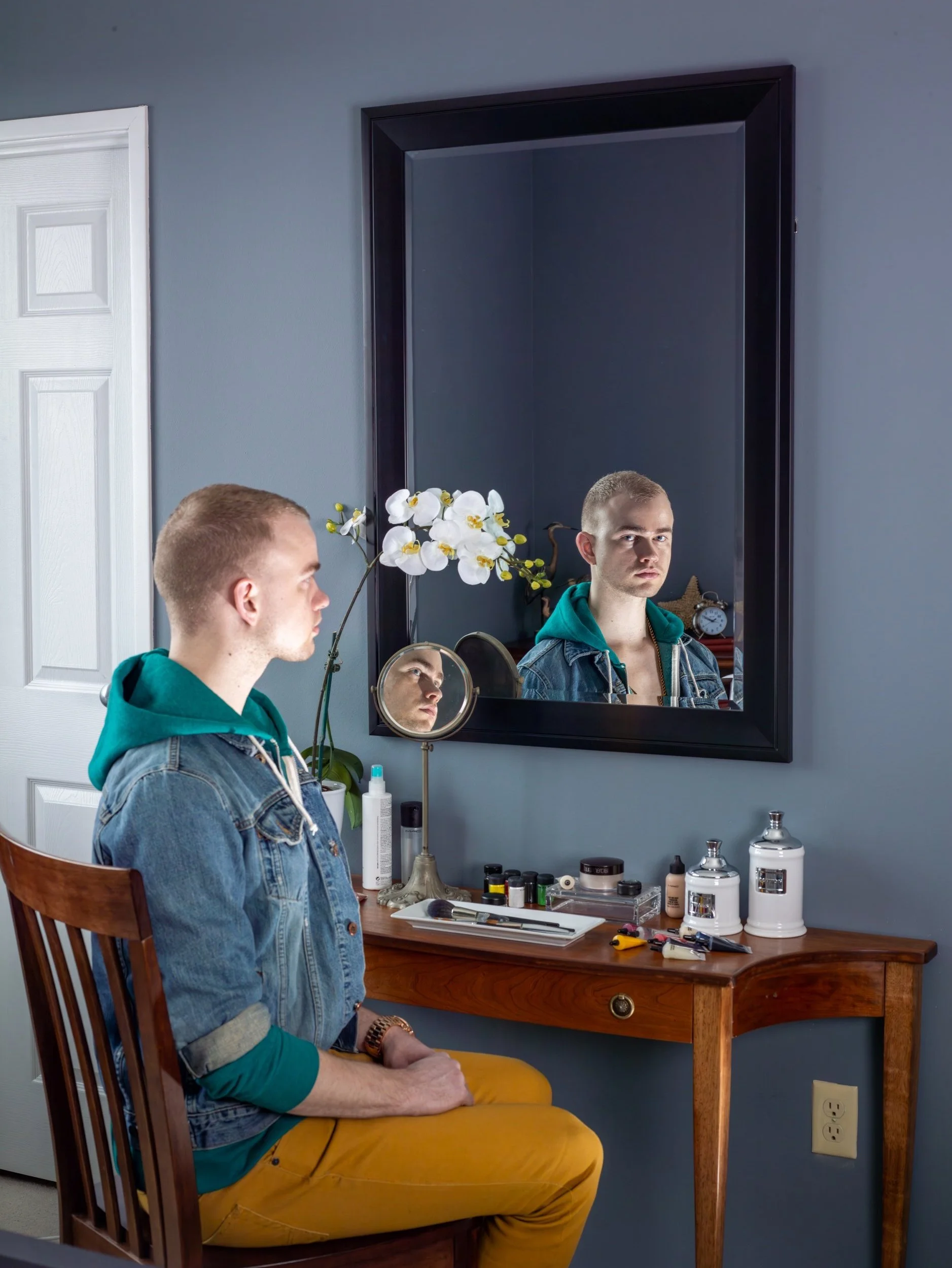 A young man sitting on a wooden chair in front of a mirror, with various grooming products on a wooden dresser, inside a room with a gray wall.