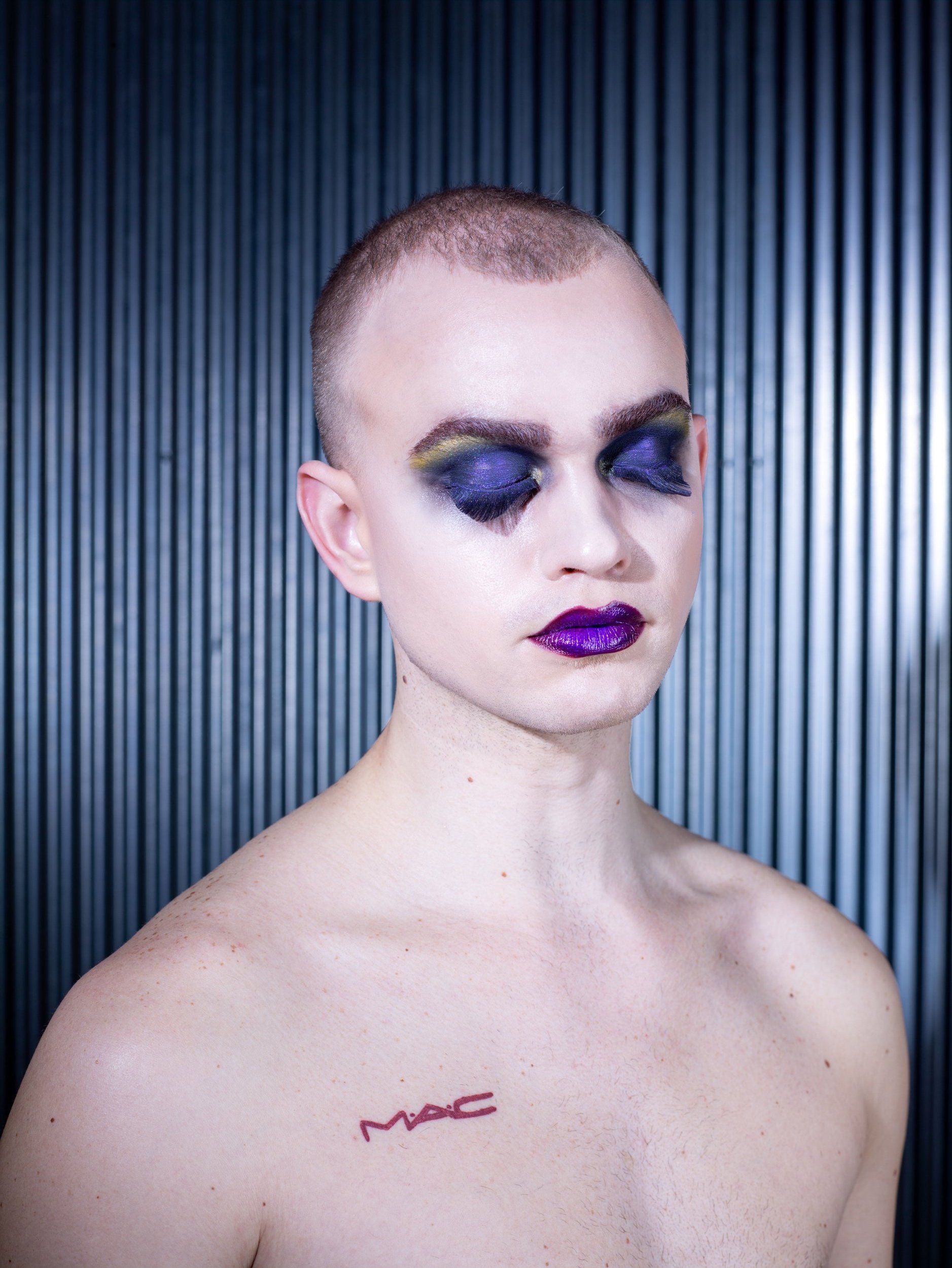A person with short hair and bold makeup featuring purple eyeshadow and dark purple lipstick, closed eyes, and a MAC tattoo on the left shoulder, standing against a metallic striped background.